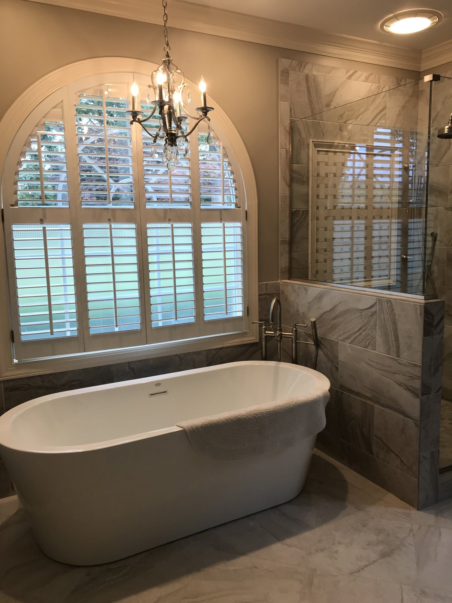 Bathroom with a white tub in front of an arched window with shutters and marble shower.