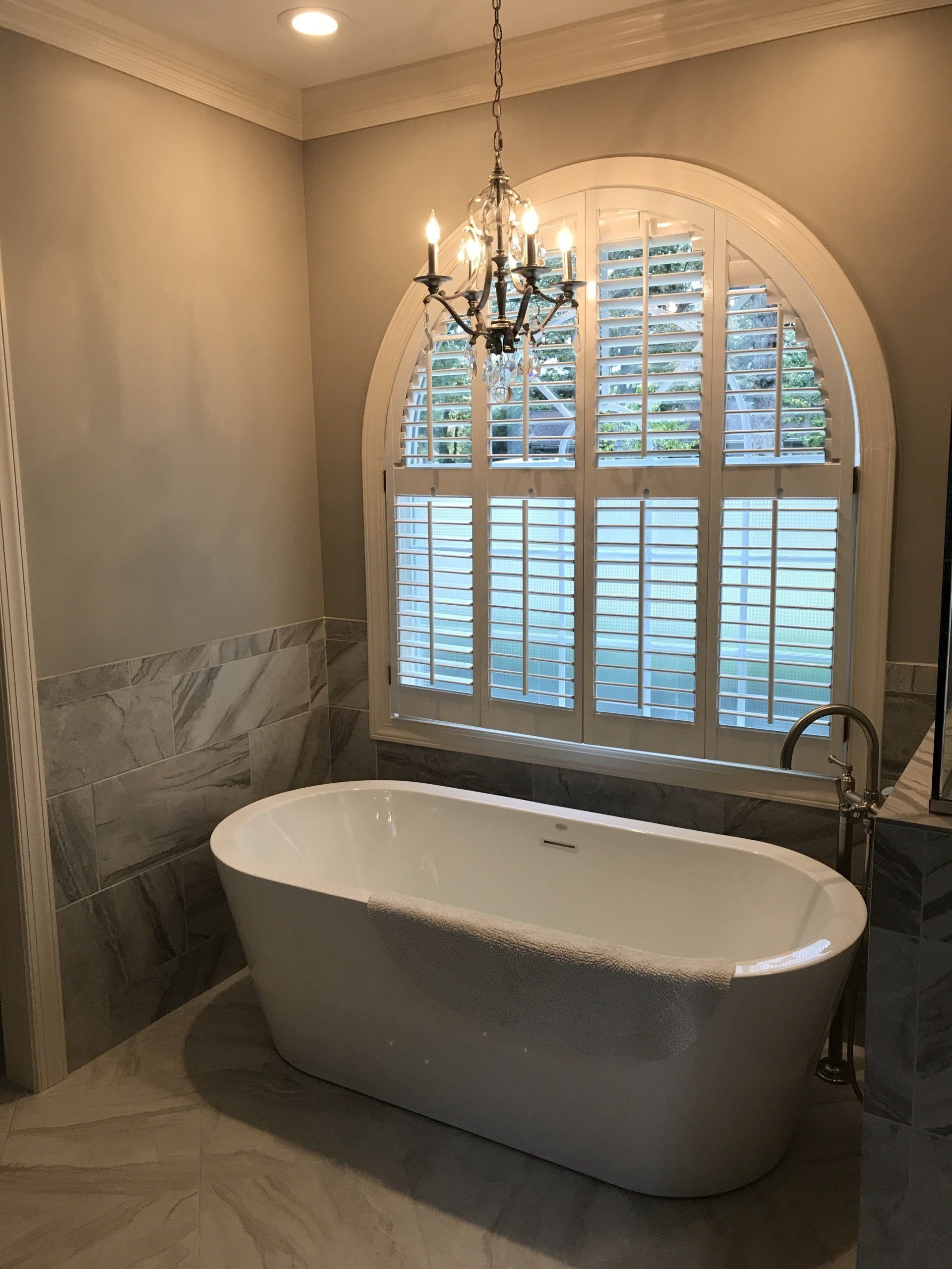 A white bathtub sits beneath an arched window with shutters, a chandelier hangs above.