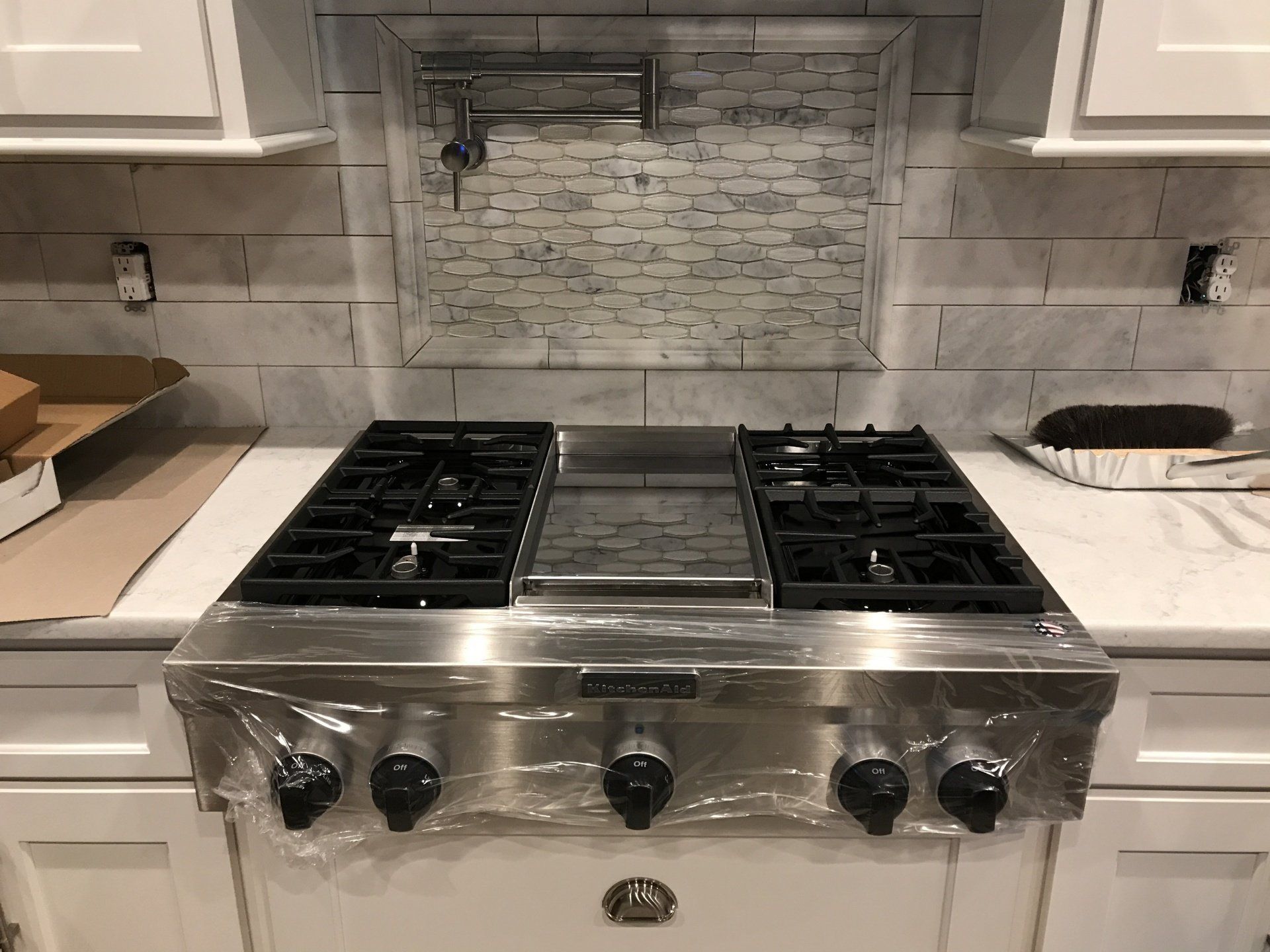 Stainless steel gas range with four burners, installed in a white kitchen, covered in plastic wrap.