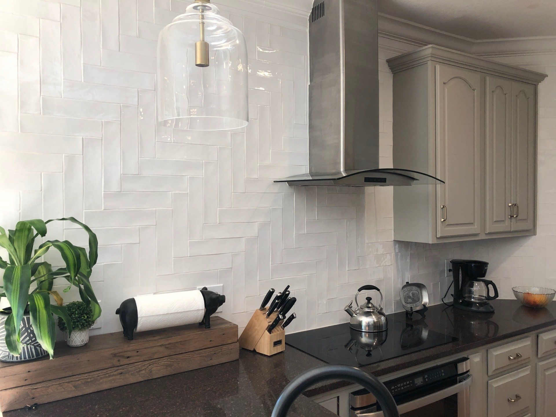 Kitchen with white herringbone tile backsplash, stainless steel range hood, grey cabinets, and dark countertop.