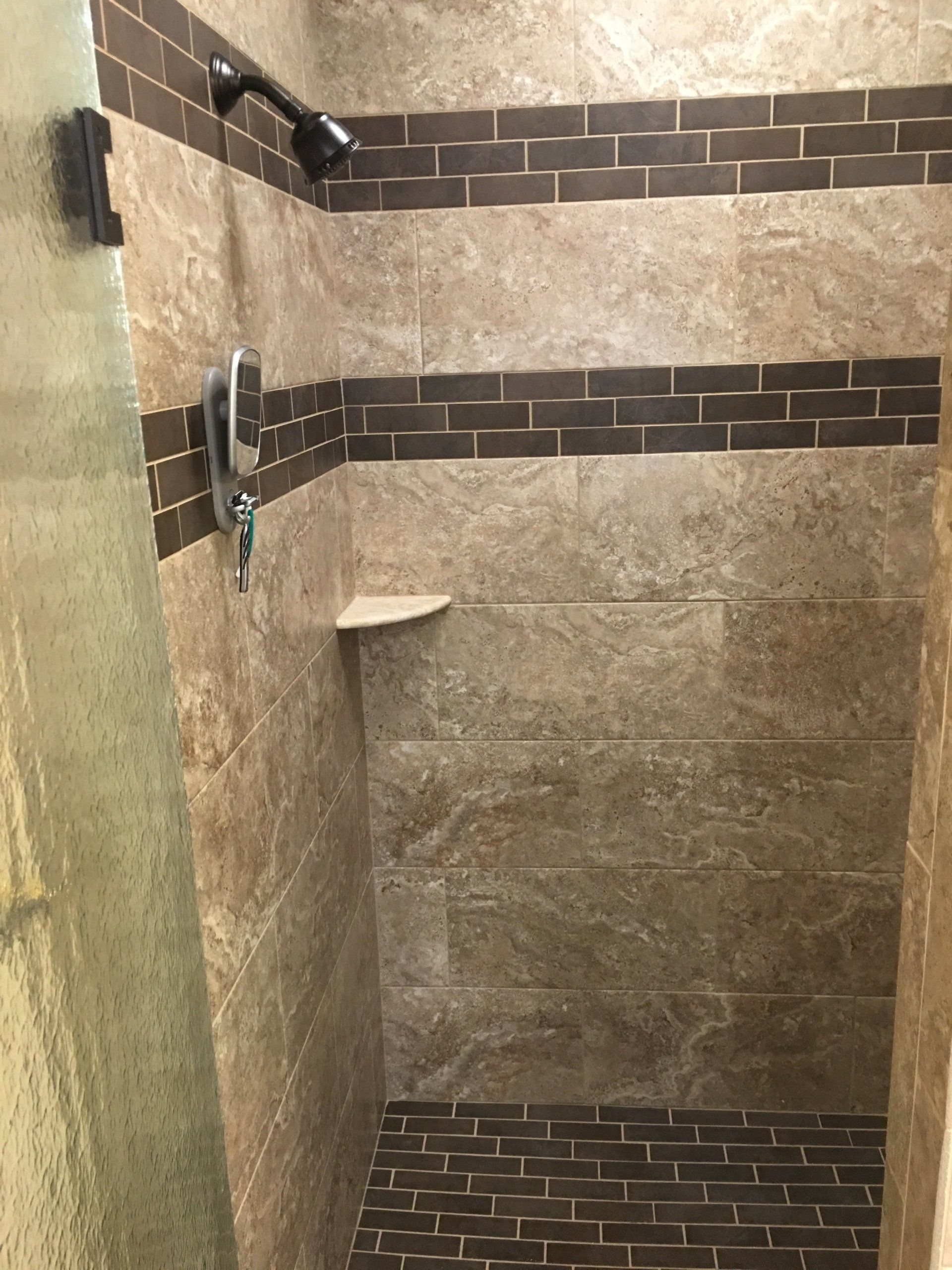 Shower with brown and tan tiles, dark accent band, built-in shelf, and glass door.