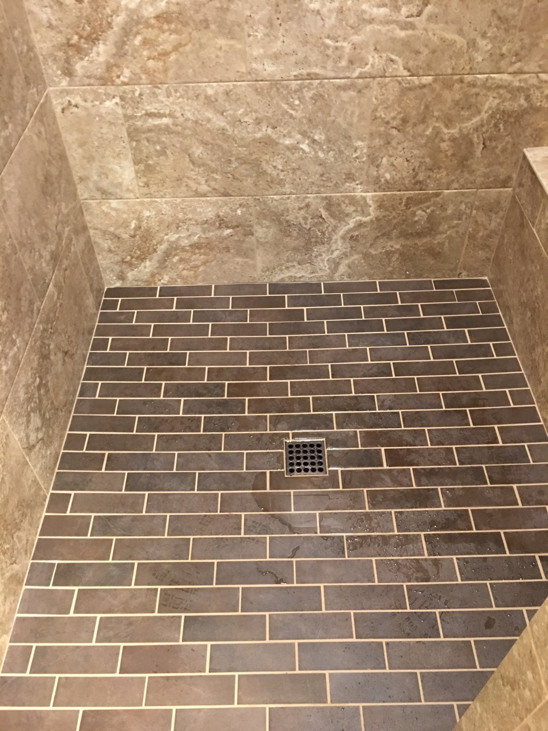 Shower with dark brick-patterned tile floor and light-colored grout. Stainless steel drain. Beige stone walls.