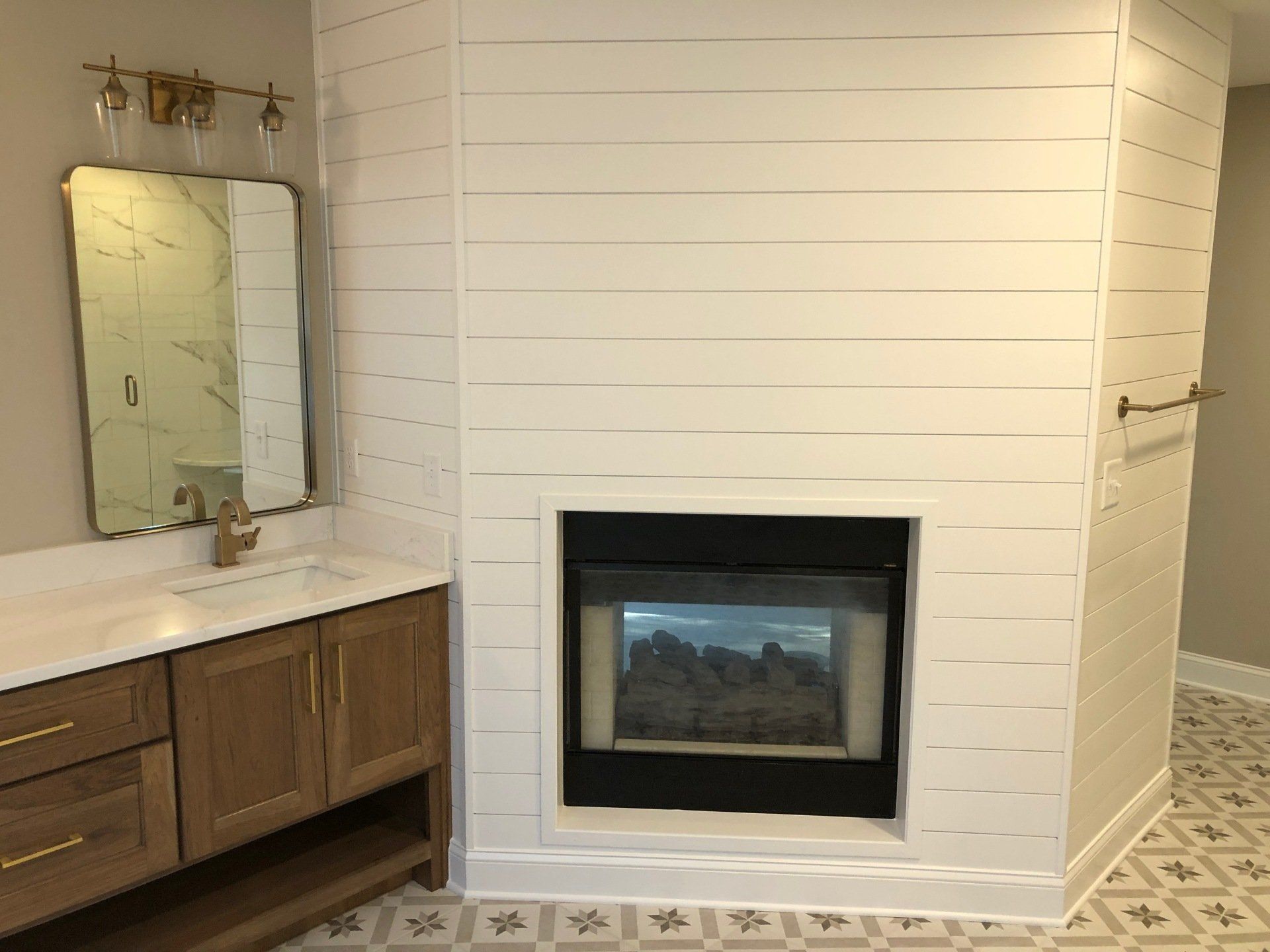 Bathroom with fireplace and vanity. White shiplap walls, wood vanity, tile floor, and gold fixtures.