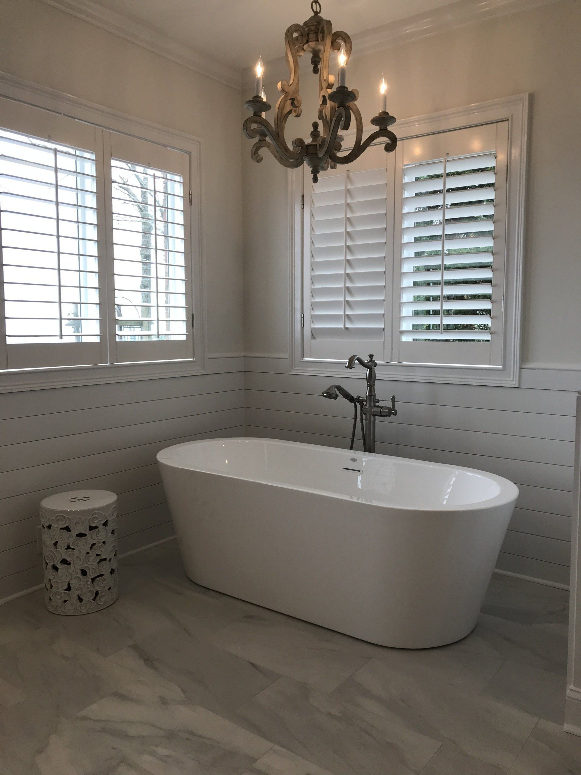 Bathroom with a white oval tub, chandelier, shutters, and patterned tile.