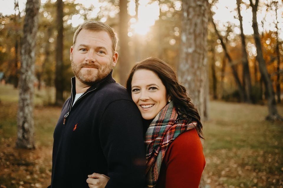 Couple smiling in a park; man in navy sweater, woman with plaid scarf, fall foliage in background.