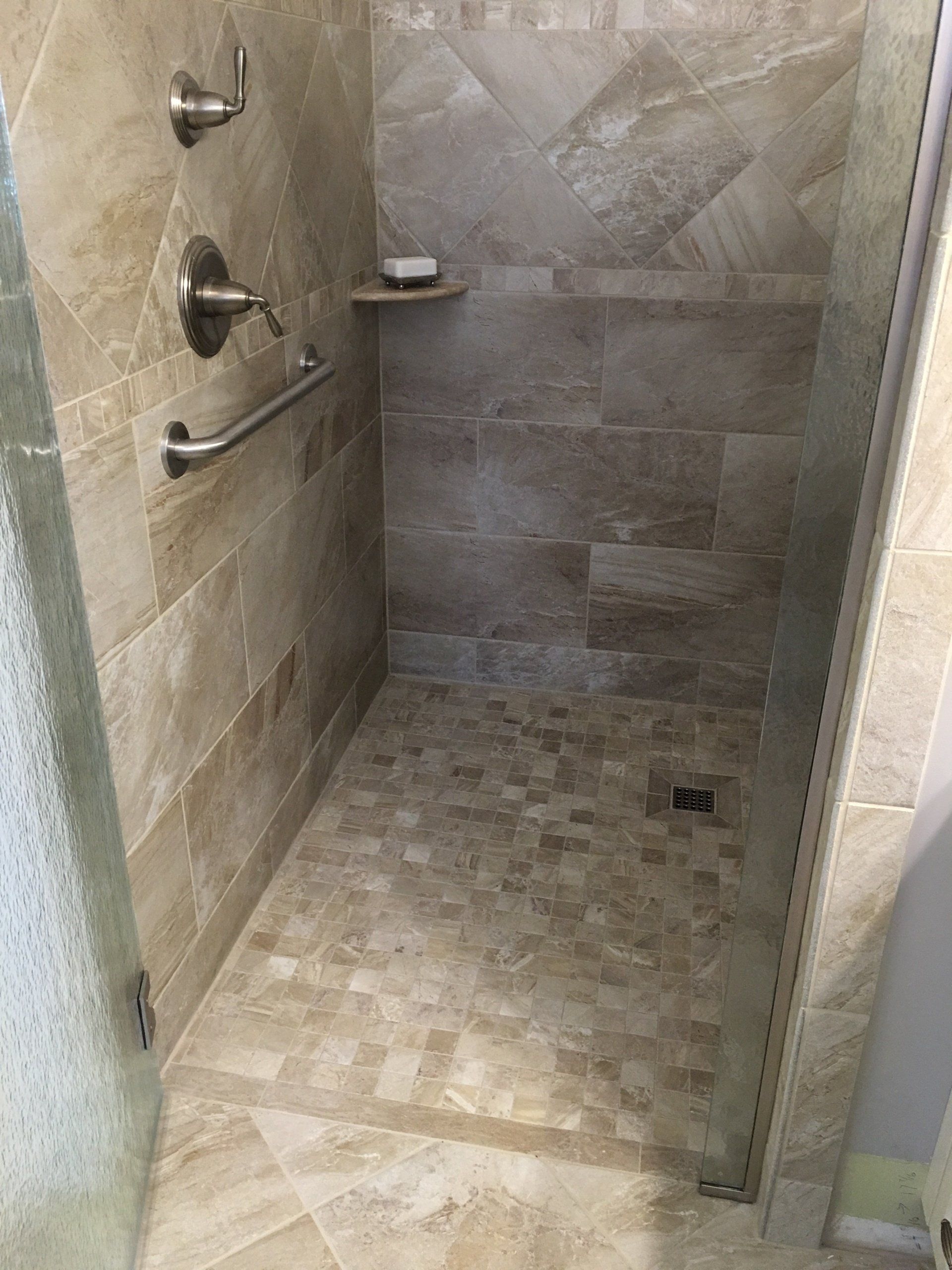 Shower with beige tile walls and floor, grab bar, soap shelf, and glass door.