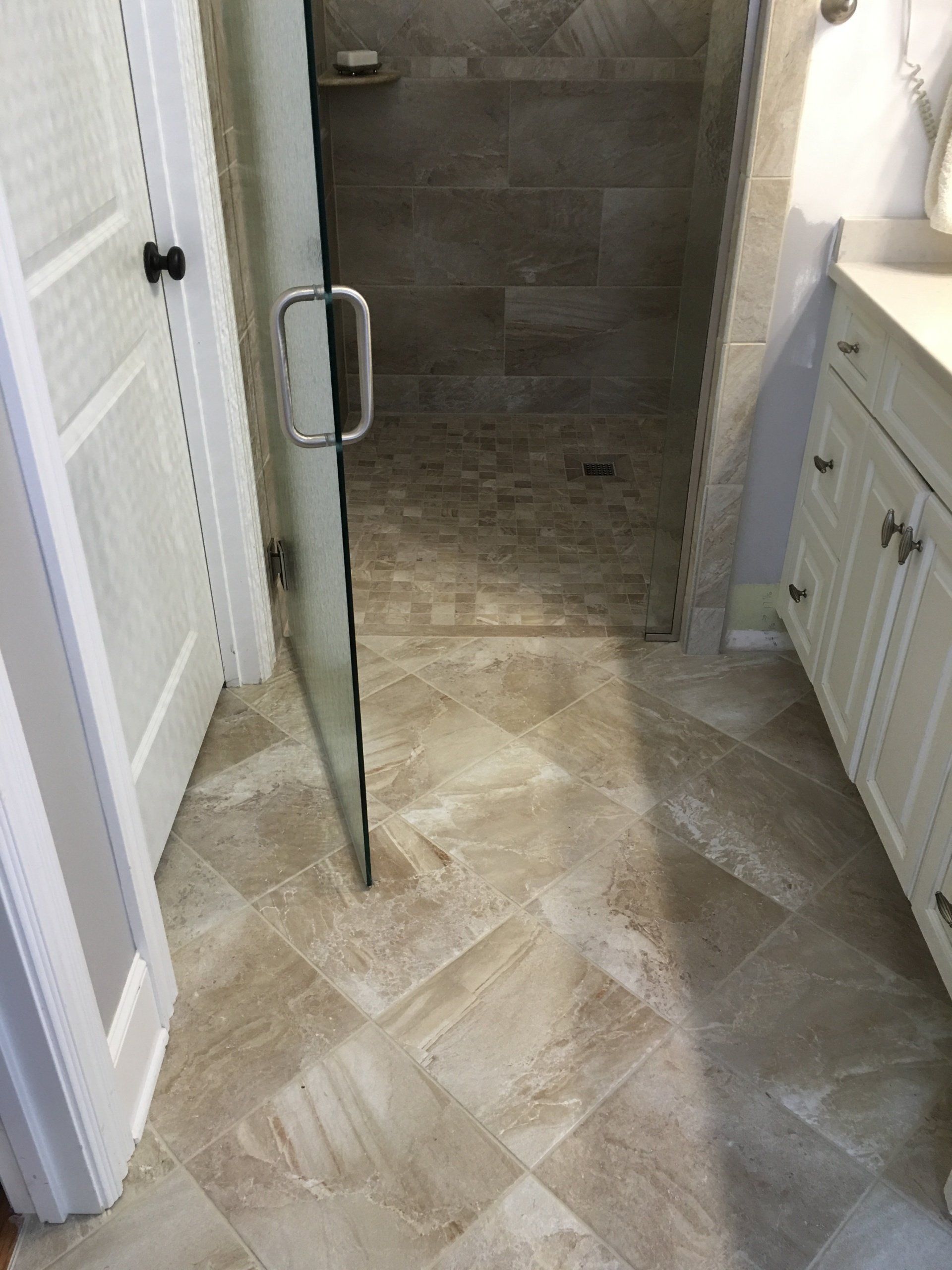 Bathroom with tile floor and shower, open door on the left, white cabinets and sink on right.