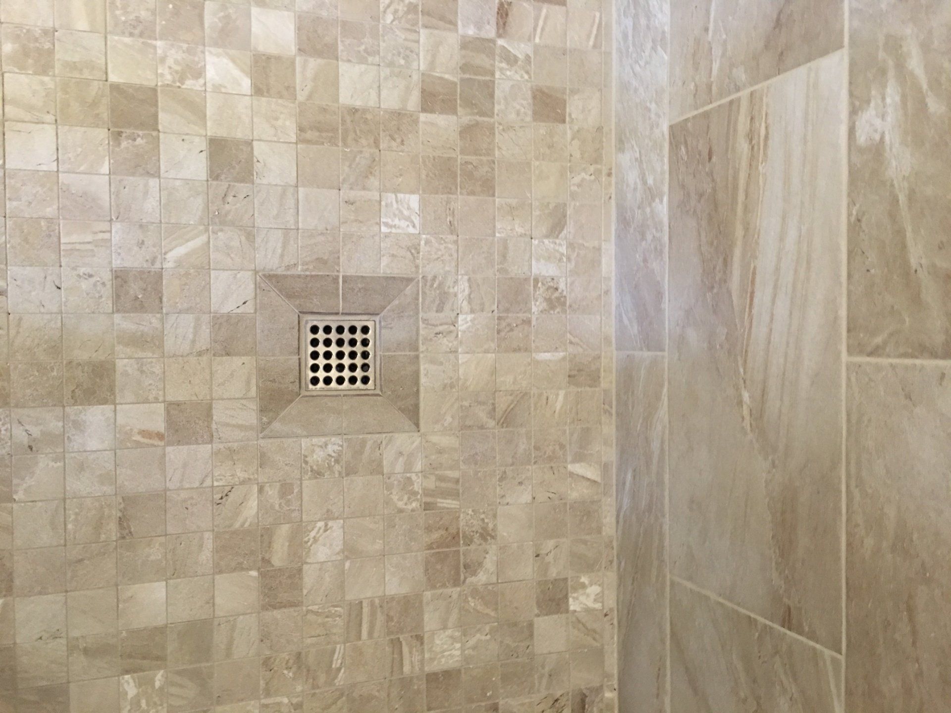 Shower with beige square and rectangular tiles, a central drain, and marble-like walls.