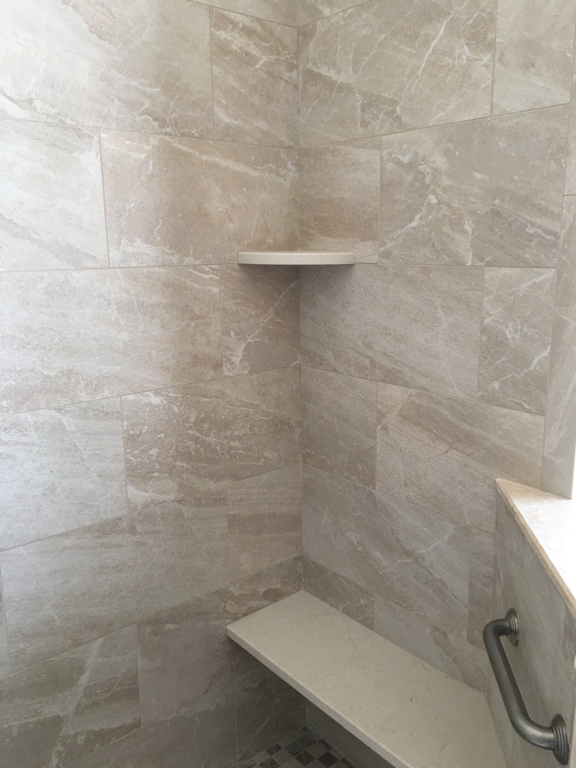 Shower with beige tile, built-in corner shelf, and seat. Silver grab bar on the right.