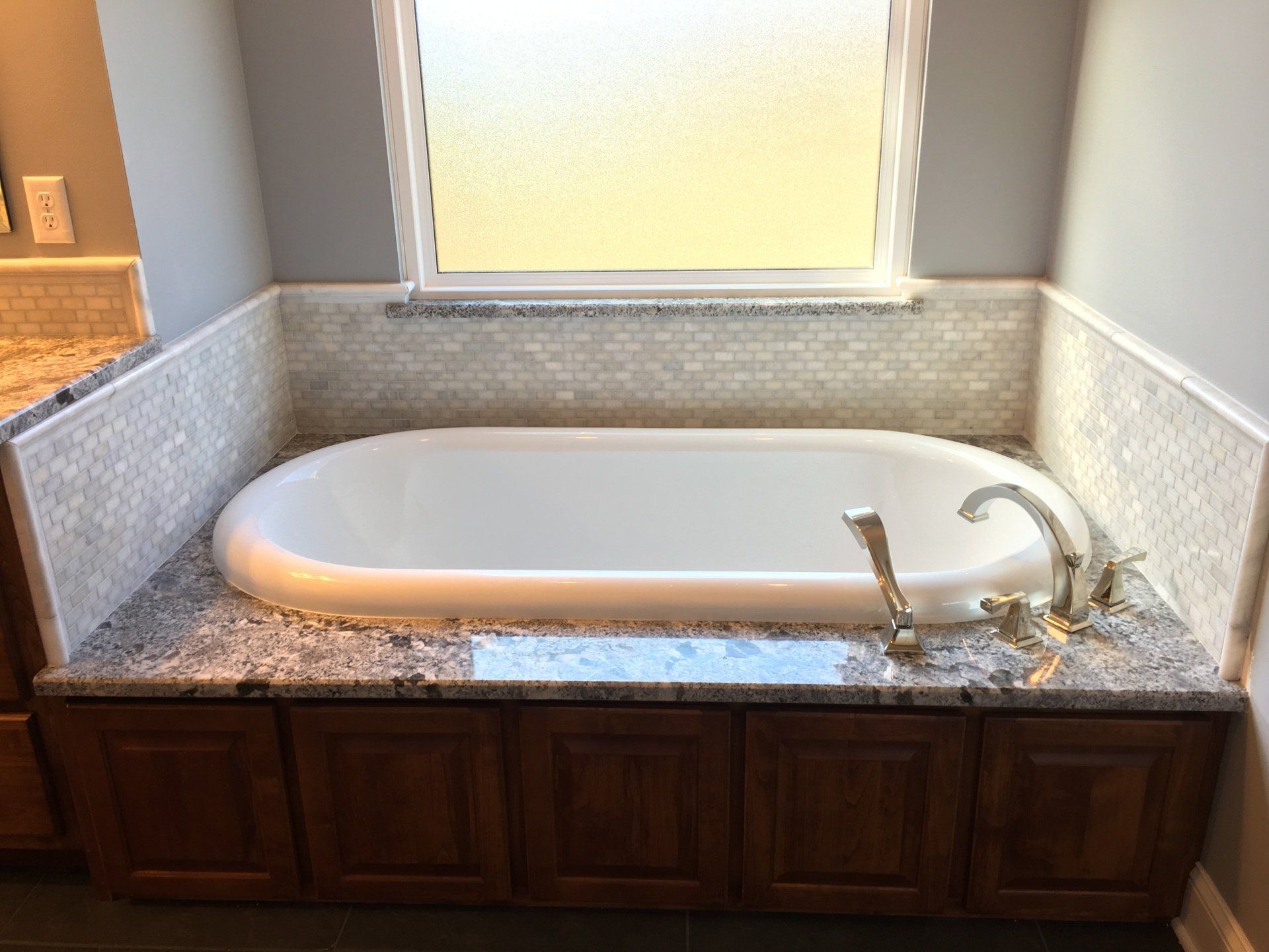 Oval bathtub set into a granite countertop with cabinets beneath and a window above, gray and wood tones.