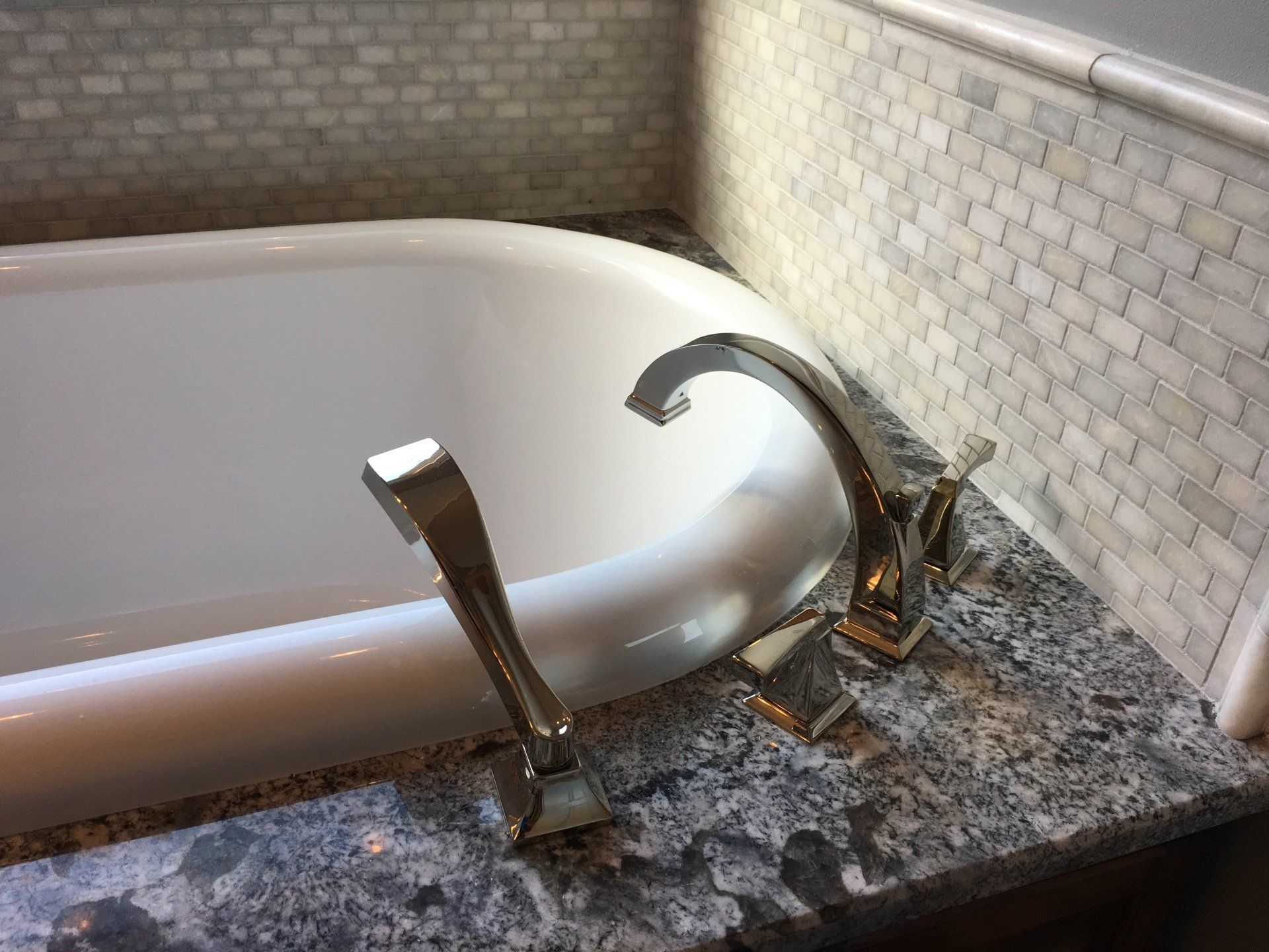Clawfoot tub with silver faucet on a granite countertop, surrounded by brick-like tile.