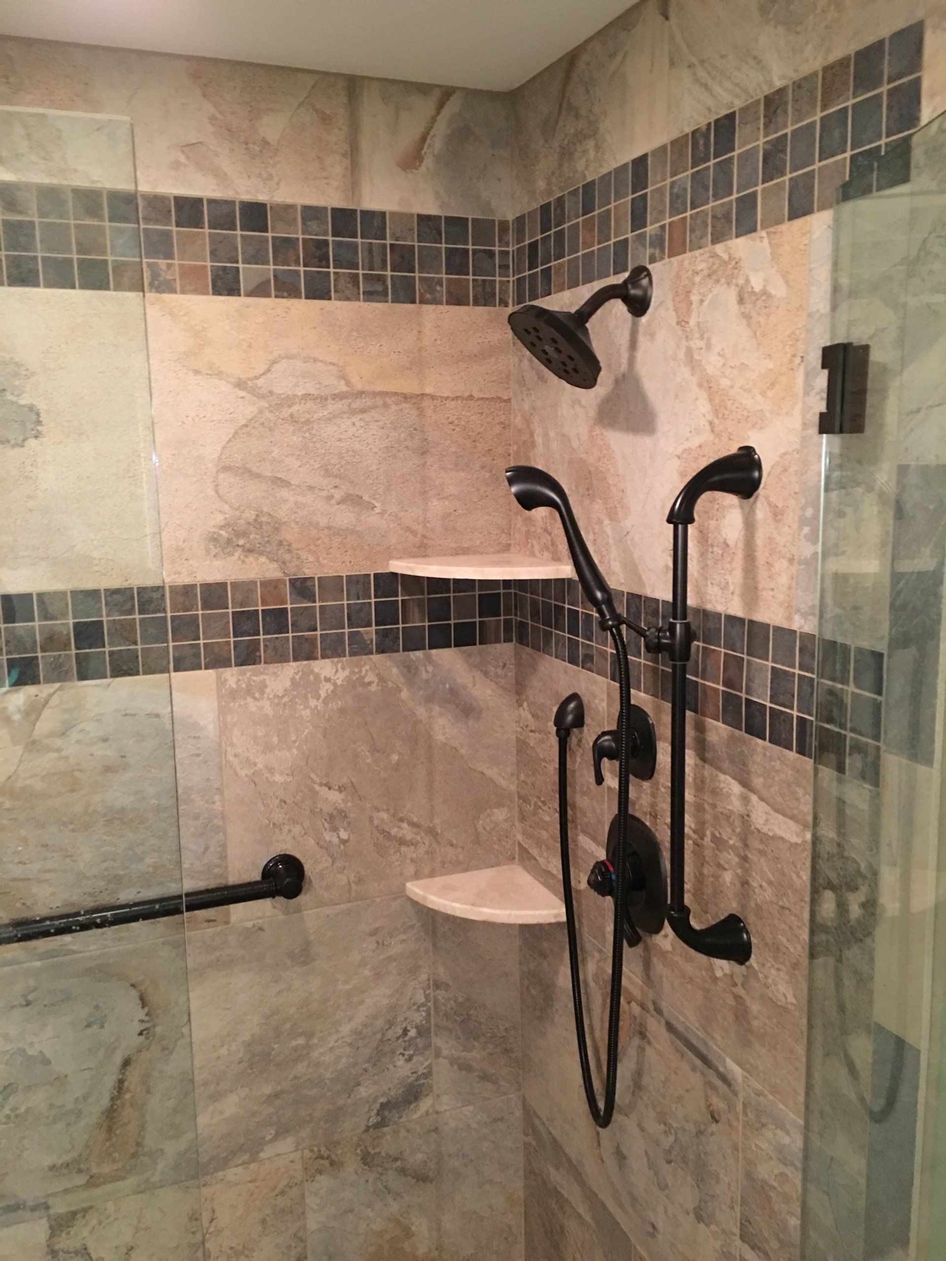 Shower stall with tan and brown stone tiles, shelves, and black fixtures, including a grab bar and hand shower.