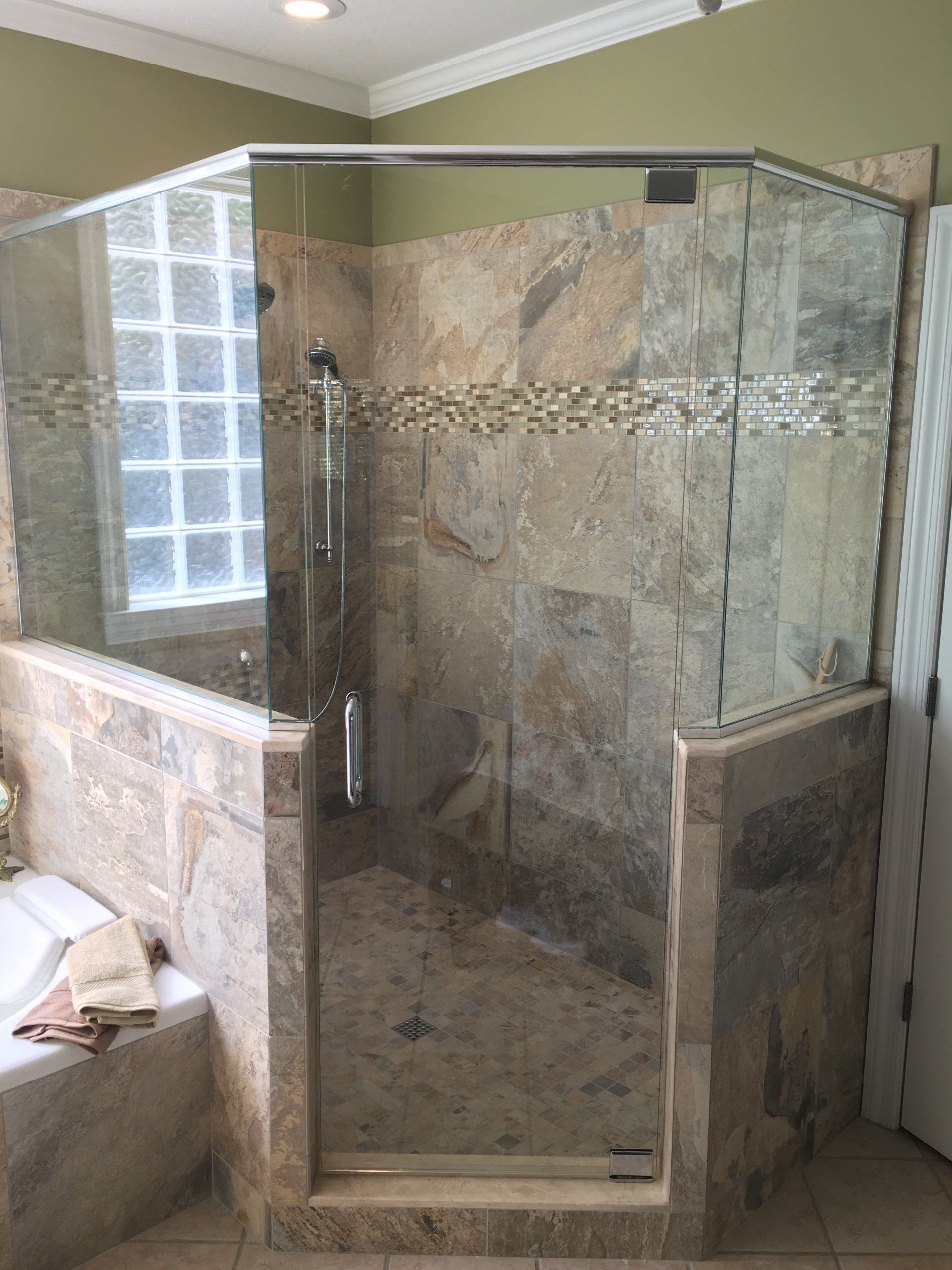 A walk-in shower with glass panels, stone tiles, and a window, next to a bathtub.