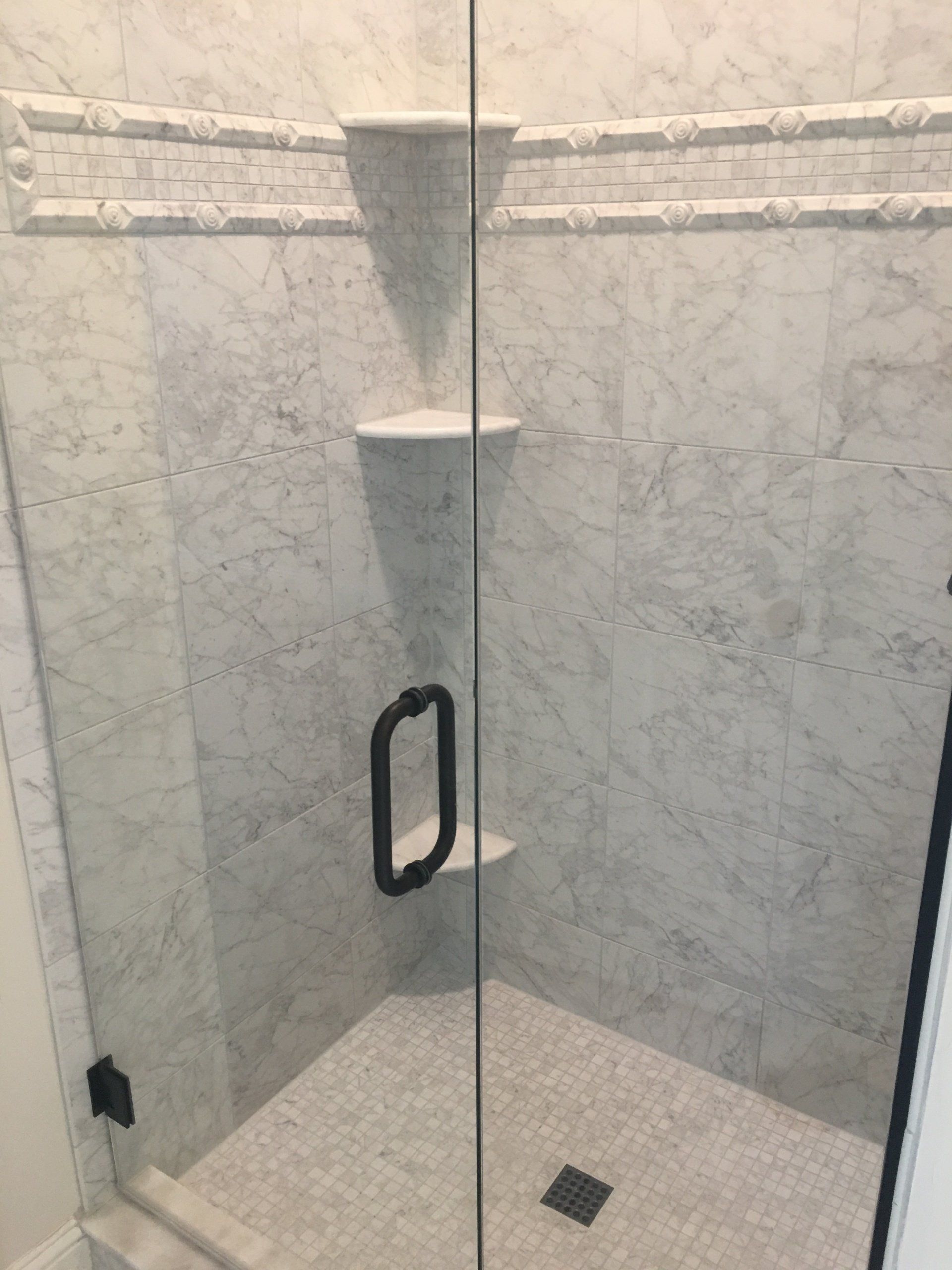 Marble-tiled shower with glass door and black hardware. Two shelves, a handle, and a drain are visible.