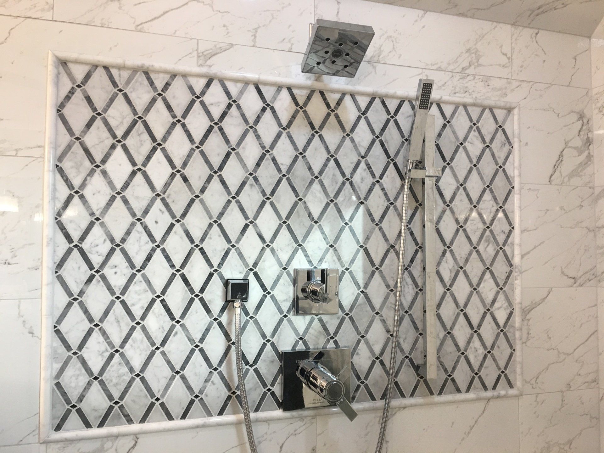 Shower wall with a diamond-patterned mosaic tile inset framed by marble tiles and a silver shower head and fixtures.