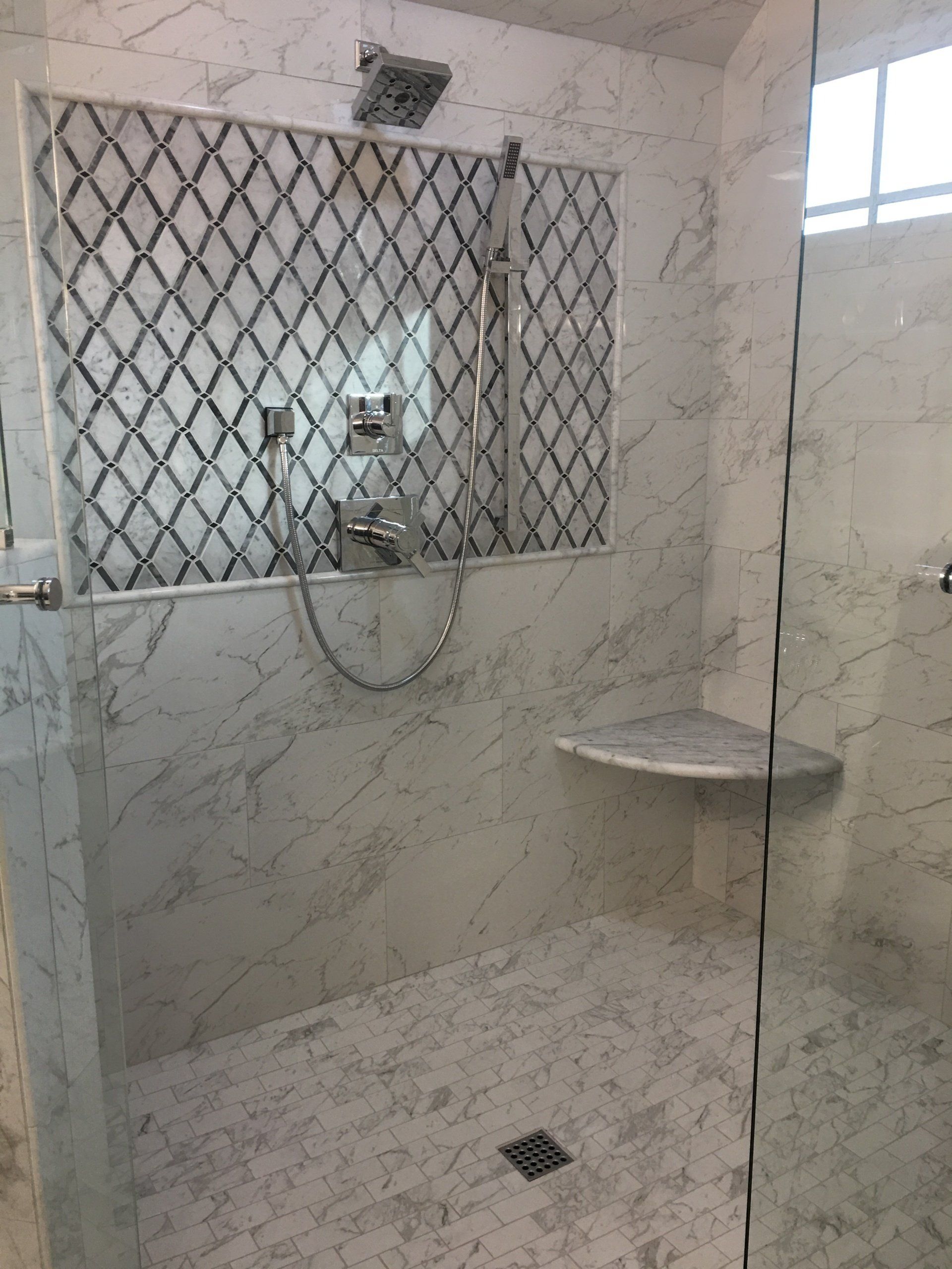 A white marble shower with a decorative tile accent, corner seat, and glass door.