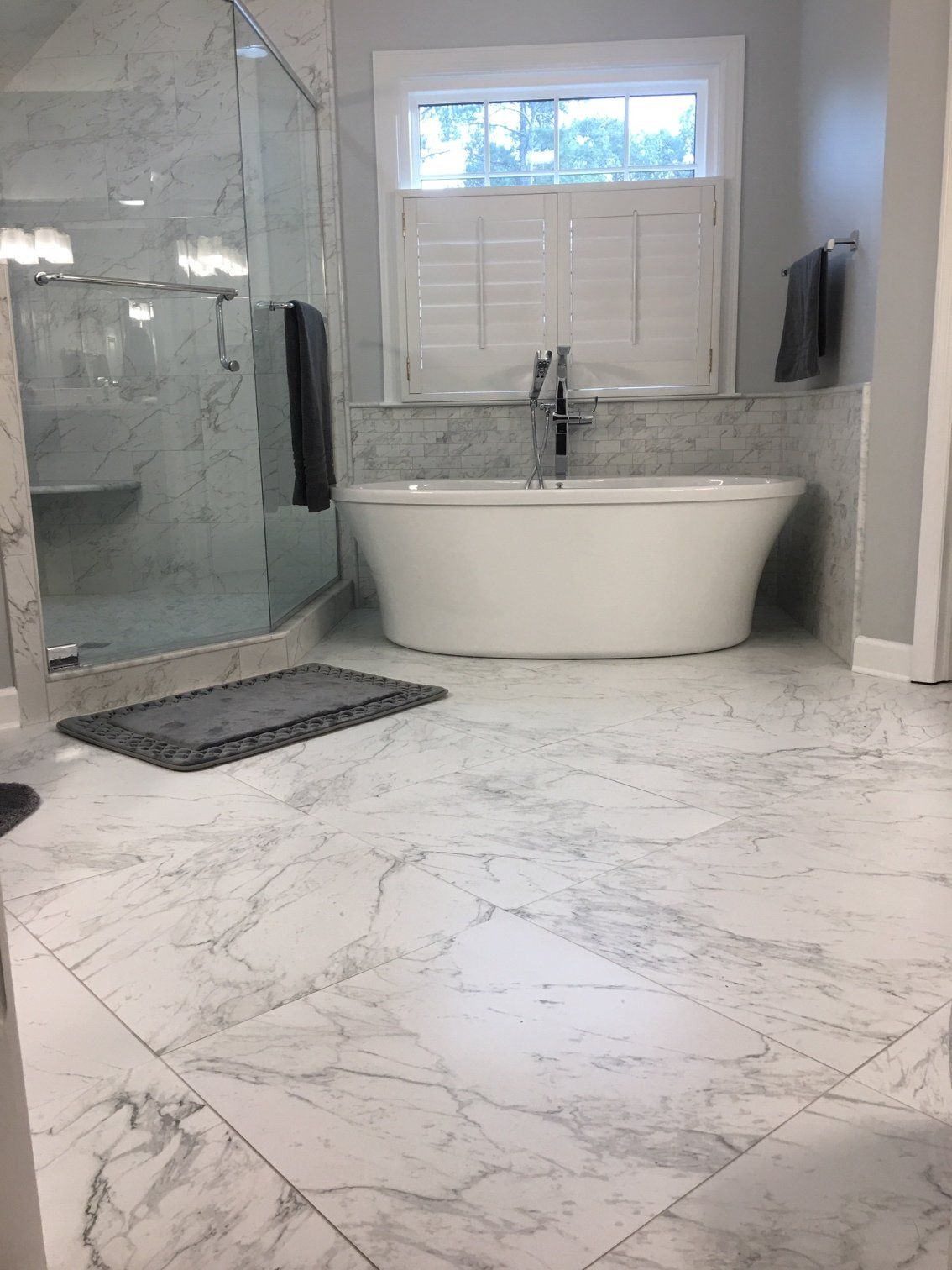 Luxurious bathroom with marble tile, freestanding tub, glass shower, and window with shutters.
