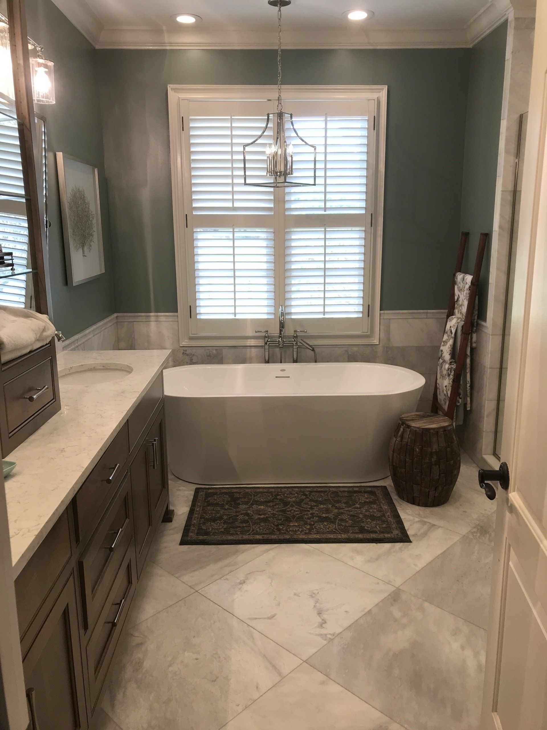 Bathroom with a white freestanding tub, blue-green walls, marble floor, and window shutters.