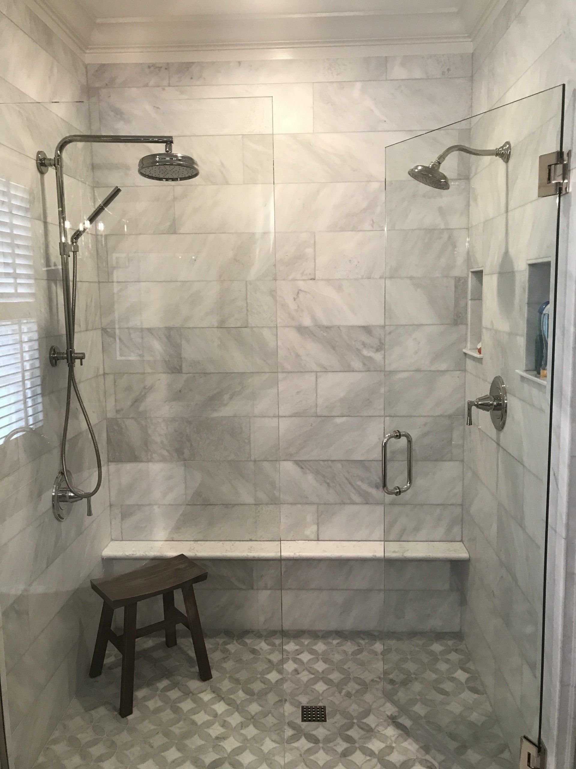 Marble-tiled shower with glass door, two showerheads, a bench, and a stool.