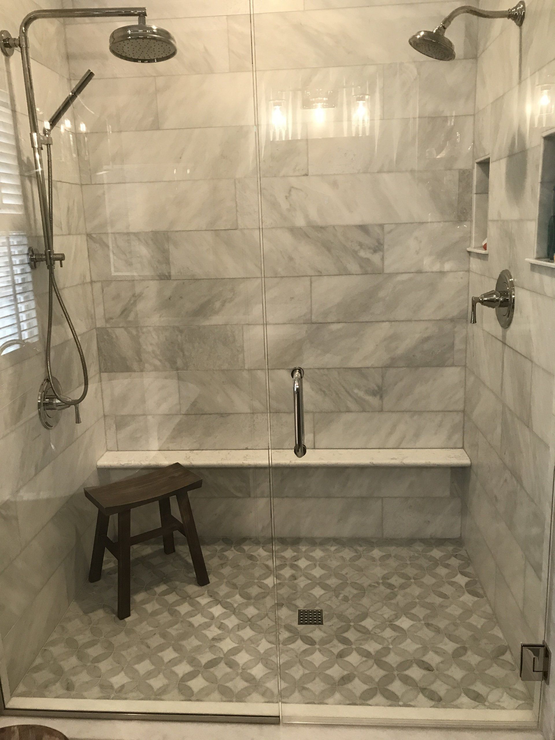 Marble tiled shower with two shower heads, a bench, and a wooden stool.