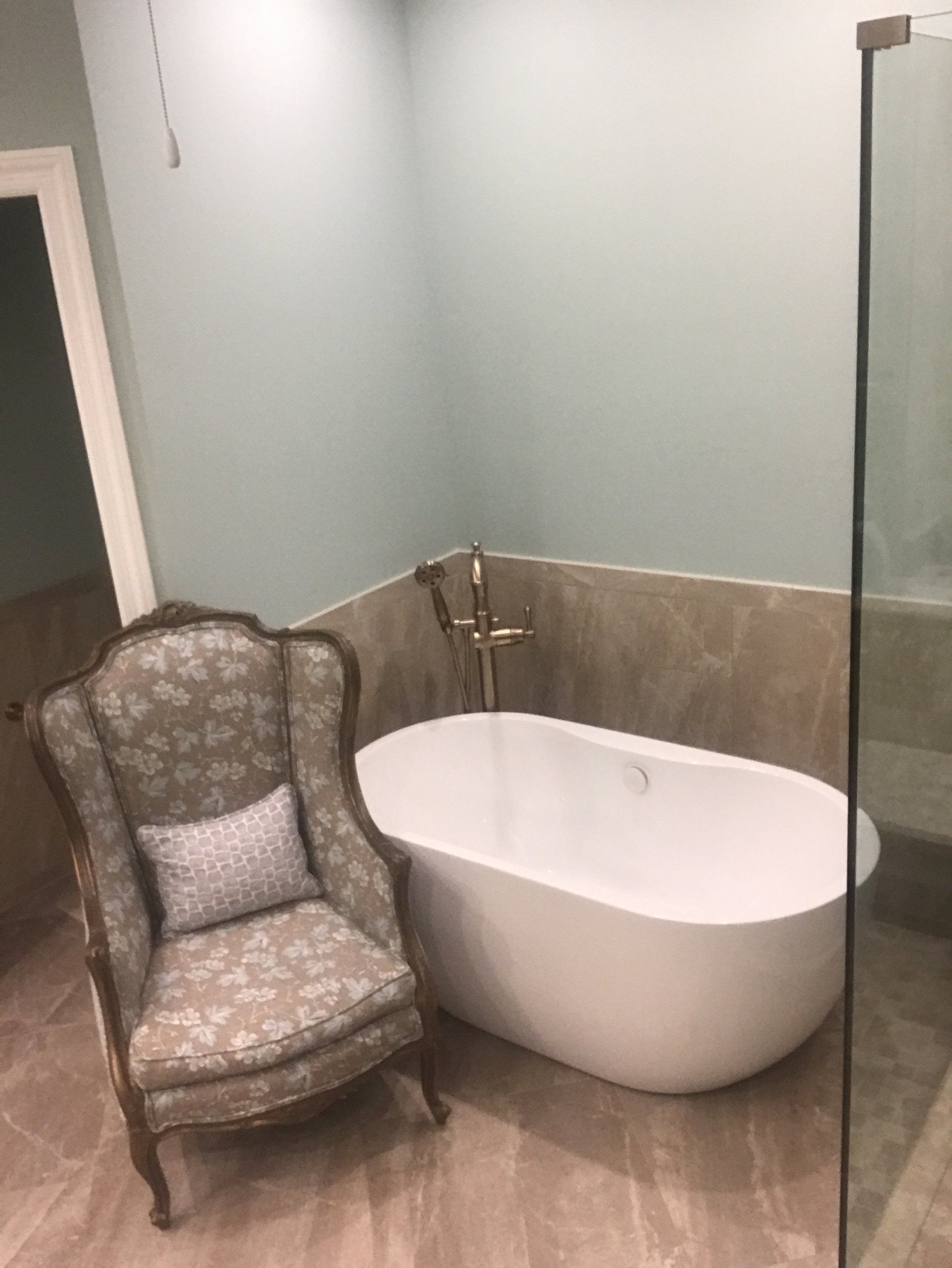 Bathroom with a white bathtub, ornate chair, and glass shower. Pale blue walls and beige tile flooring.