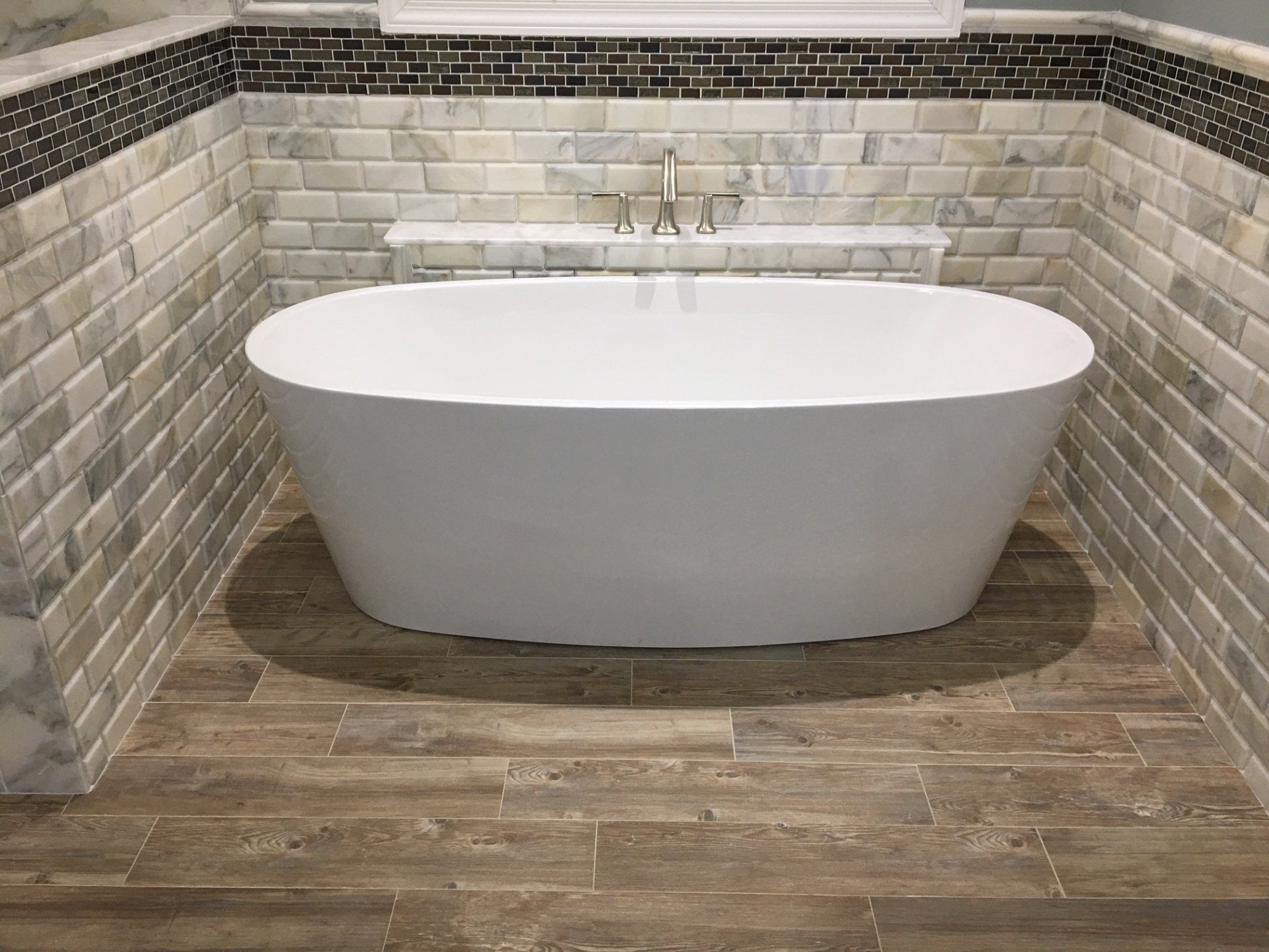 Freestanding white bathtub in a bathroom with wood-look floor tiles, marble-look wall tiles, and a shelf.