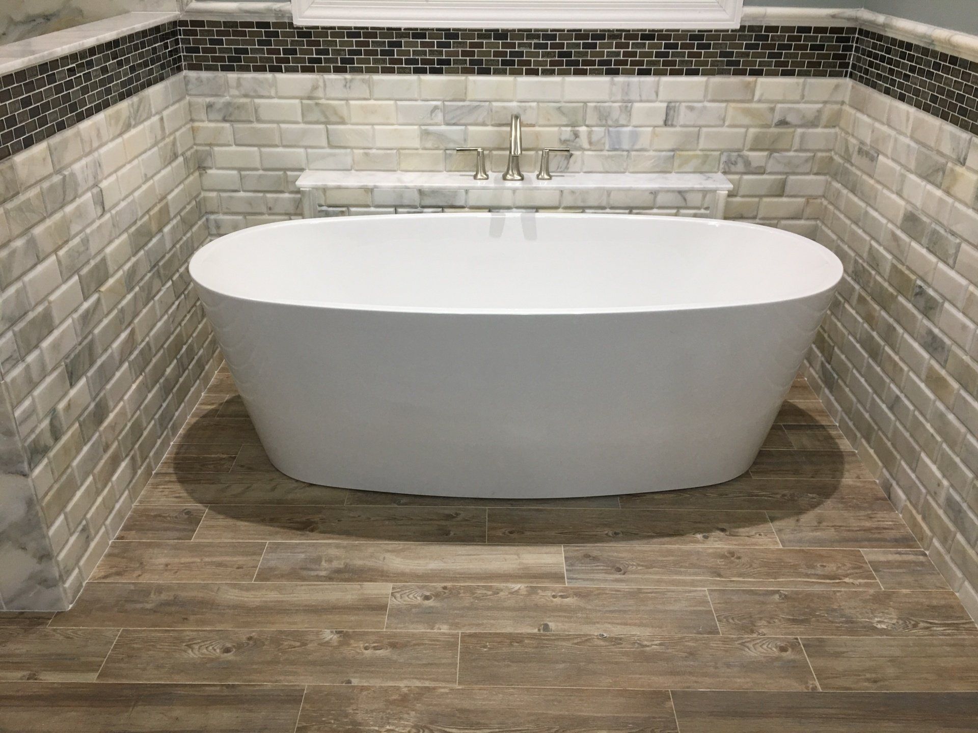 A white oval bathtub in a bathroom with marble tiles and wood-look flooring.