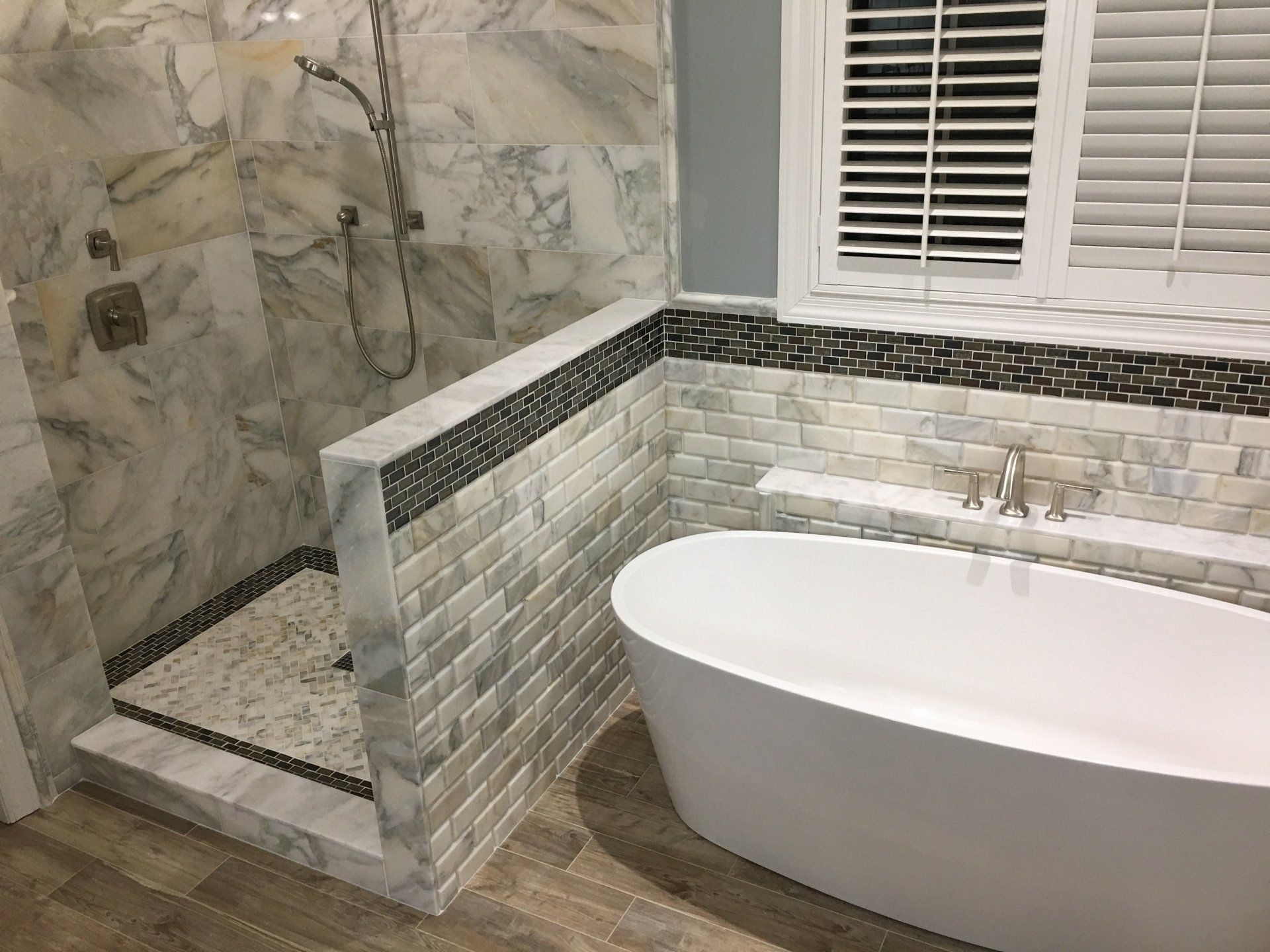 Modern bathroom with marble shower, white bathtub, and window with blinds.