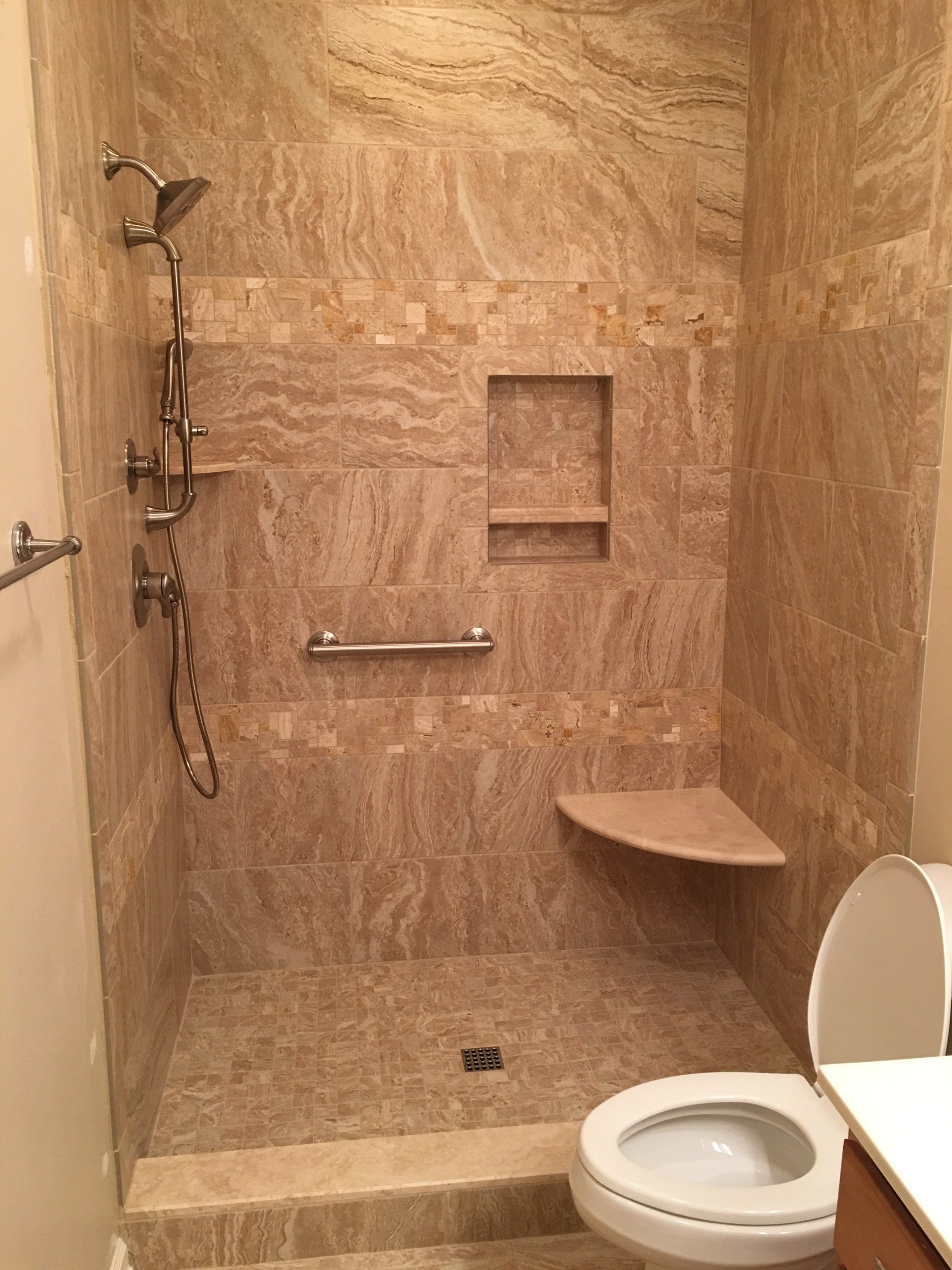 Walk-in shower with beige tile, a built-in shelf, corner seat, and grab bar, near a toilet.