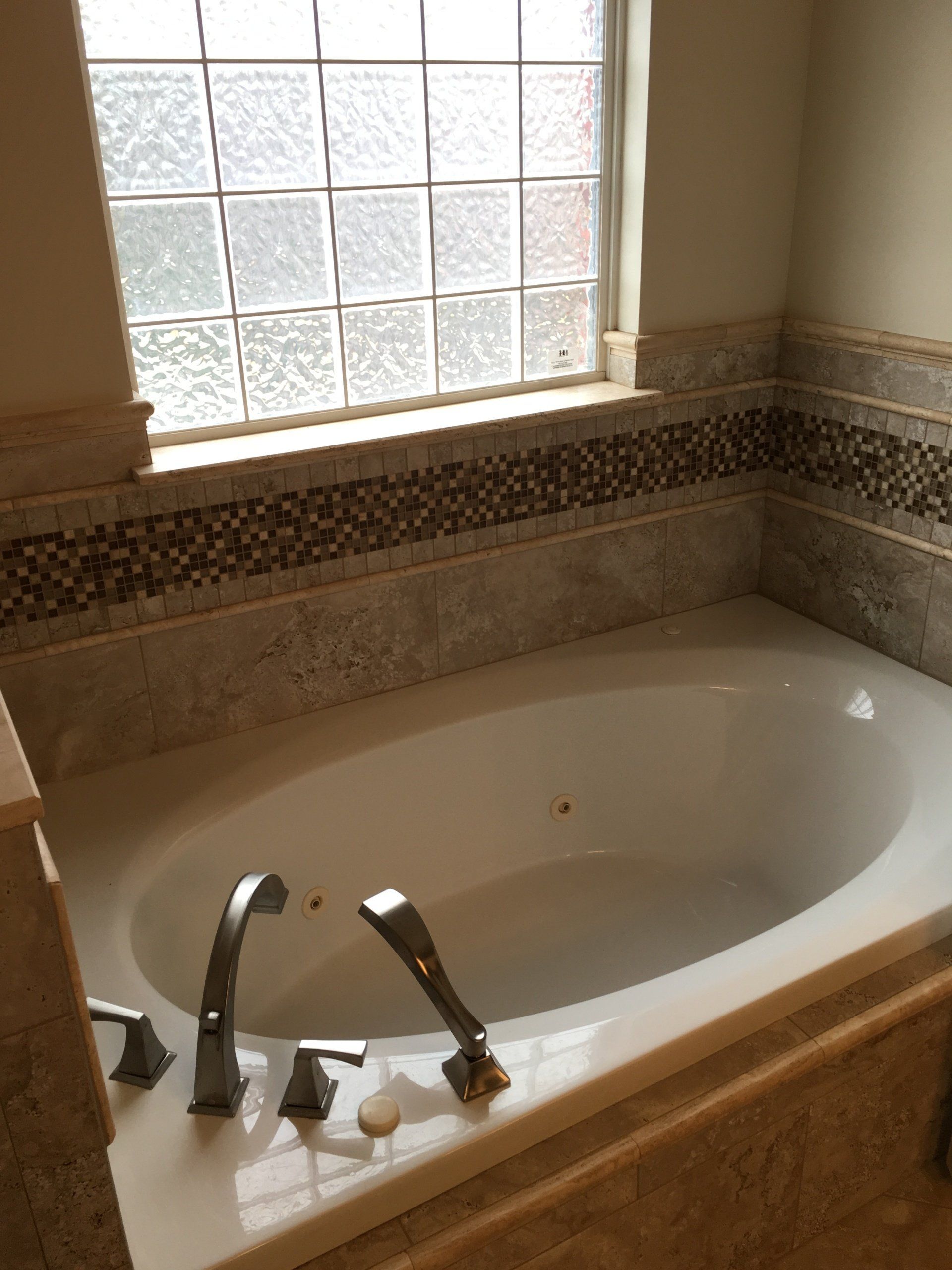 Oval bathtub with chrome fixtures, mosaic tile border, and frosted window above.