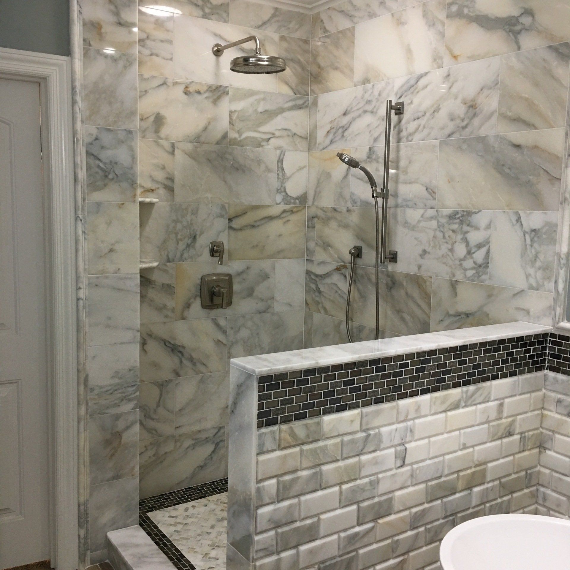Marble-tiled shower with overhead and handheld showerheads, a tiled half-wall, and a white door.