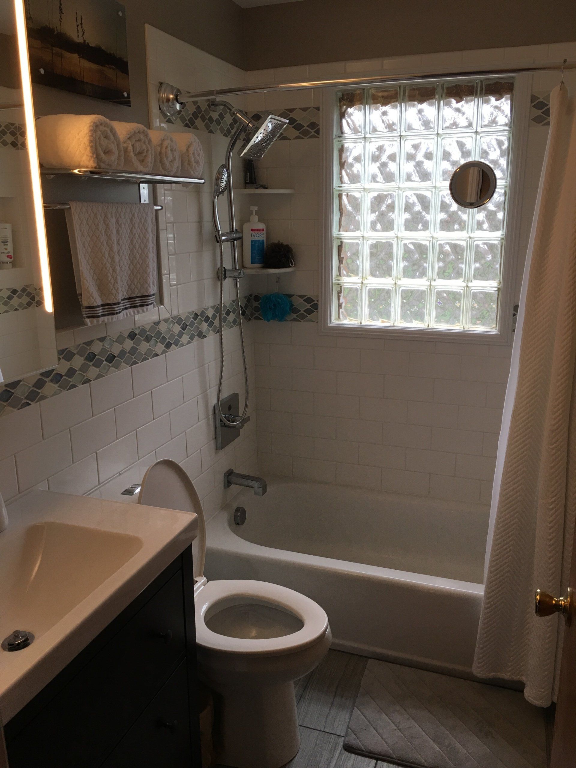 Bathroom with tub, toilet, sink. White tile, glass block window. Towels on shelf.