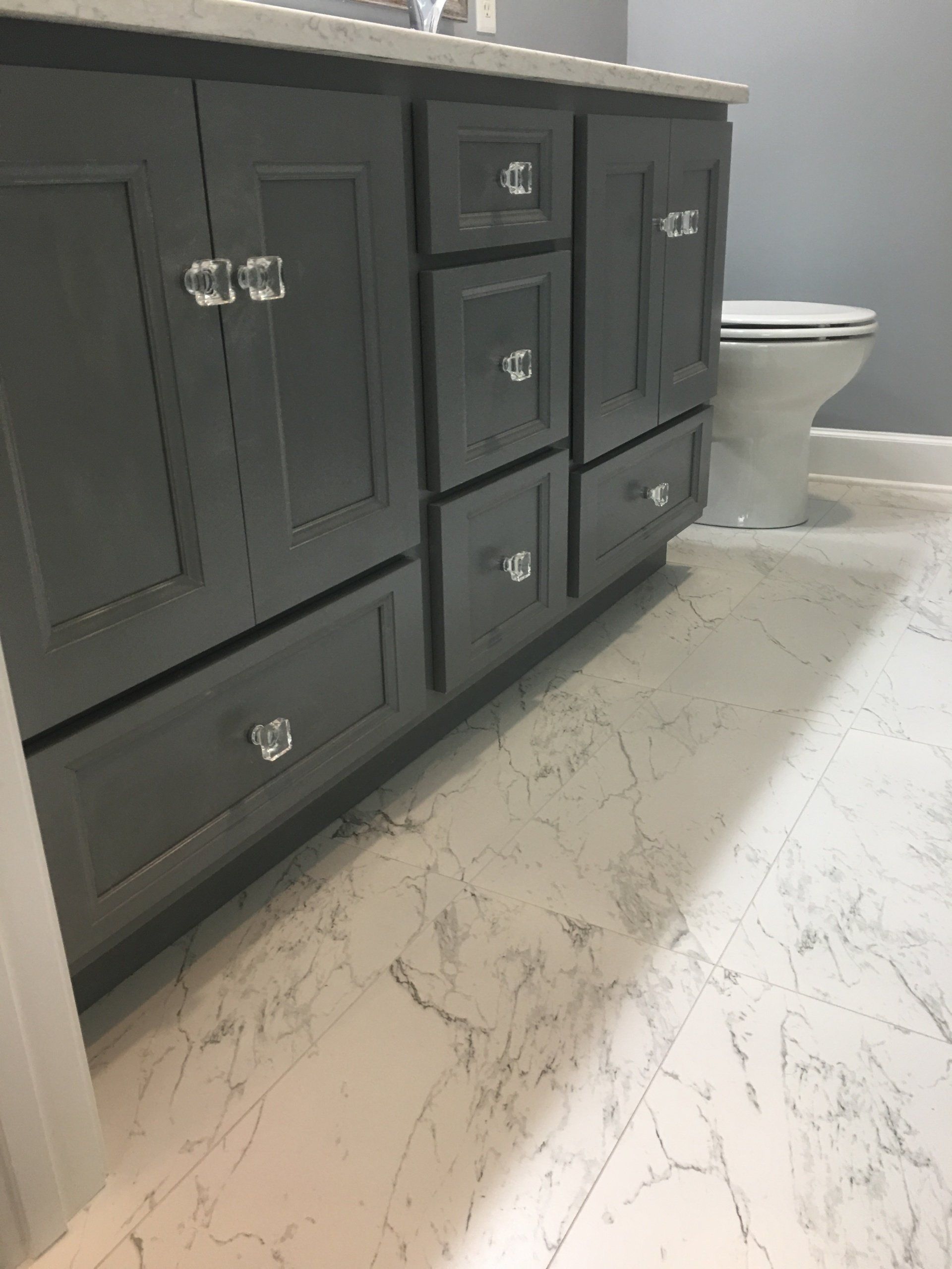 Gray bathroom vanity with crystal knobs, in front of a white marble patterned floor, toilet visible in the background.