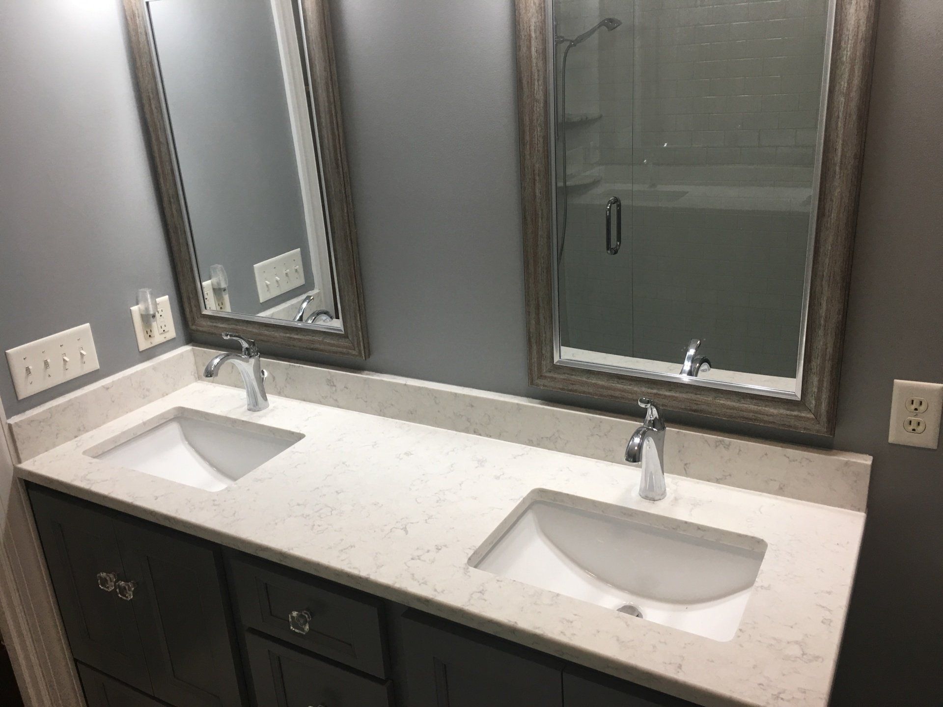 Bathroom with two sinks, mirrors, gray cabinets, and light-colored countertop.