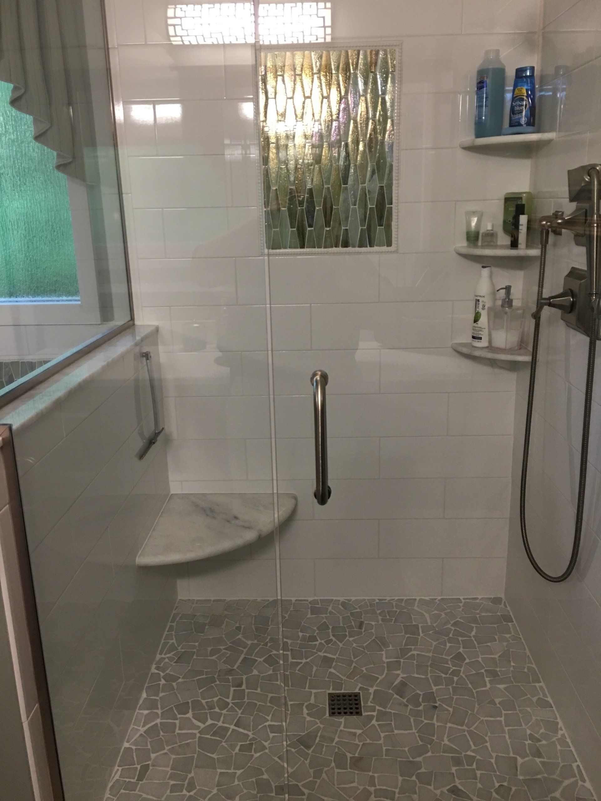 A modern white-tiled shower with a glass door, corner seat, and decorative mosaic window.