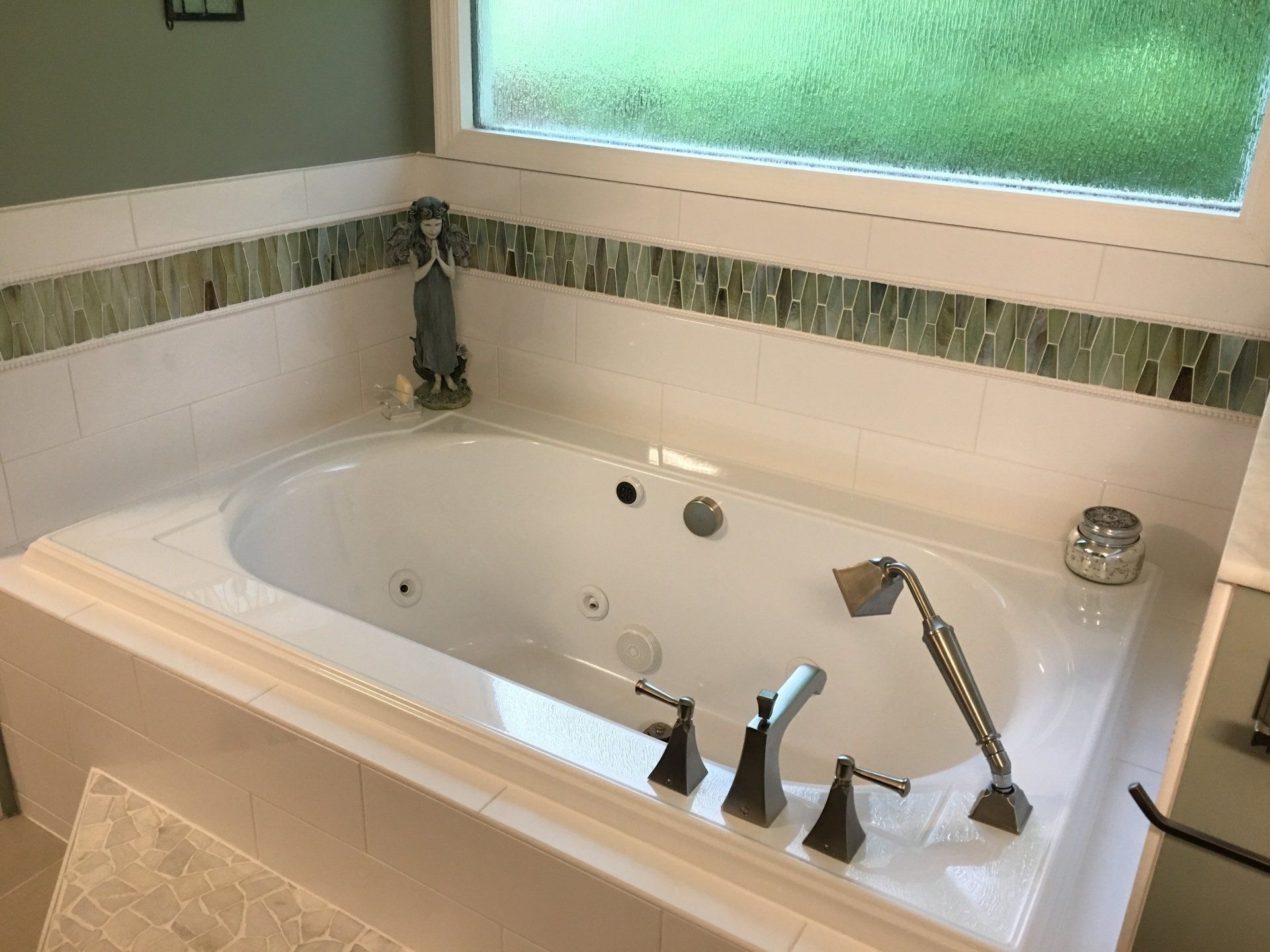 White built-in bathtub with bronze fixtures, surrounded by white tile and green glass accent tile, below a window.