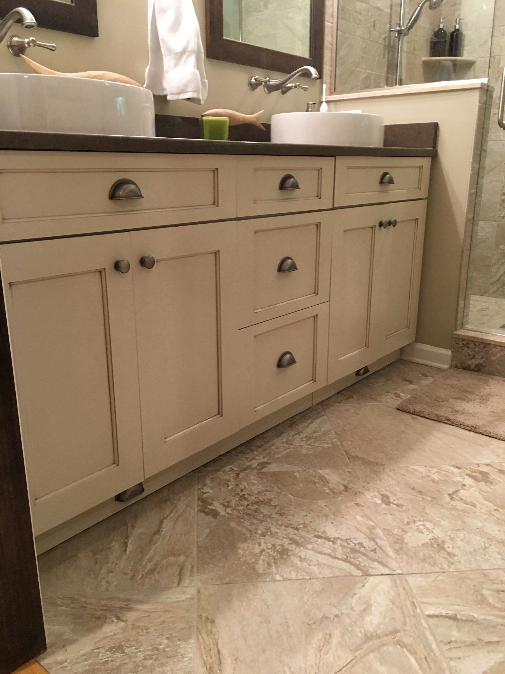 Cream-colored bathroom vanity with two sinks, dark countertop, and metal hardware, set on patterned tile flooring.