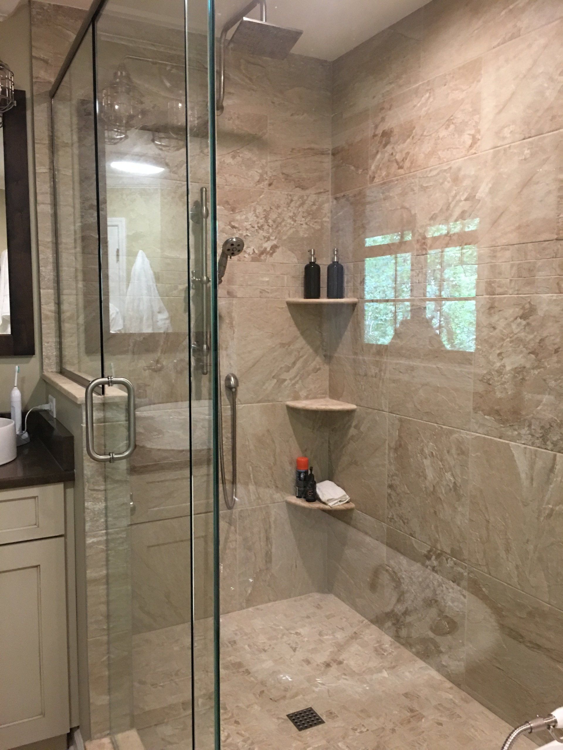Glass-enclosed shower with tan tile walls and stone floor, built-in corner shelves, and a grab bar.