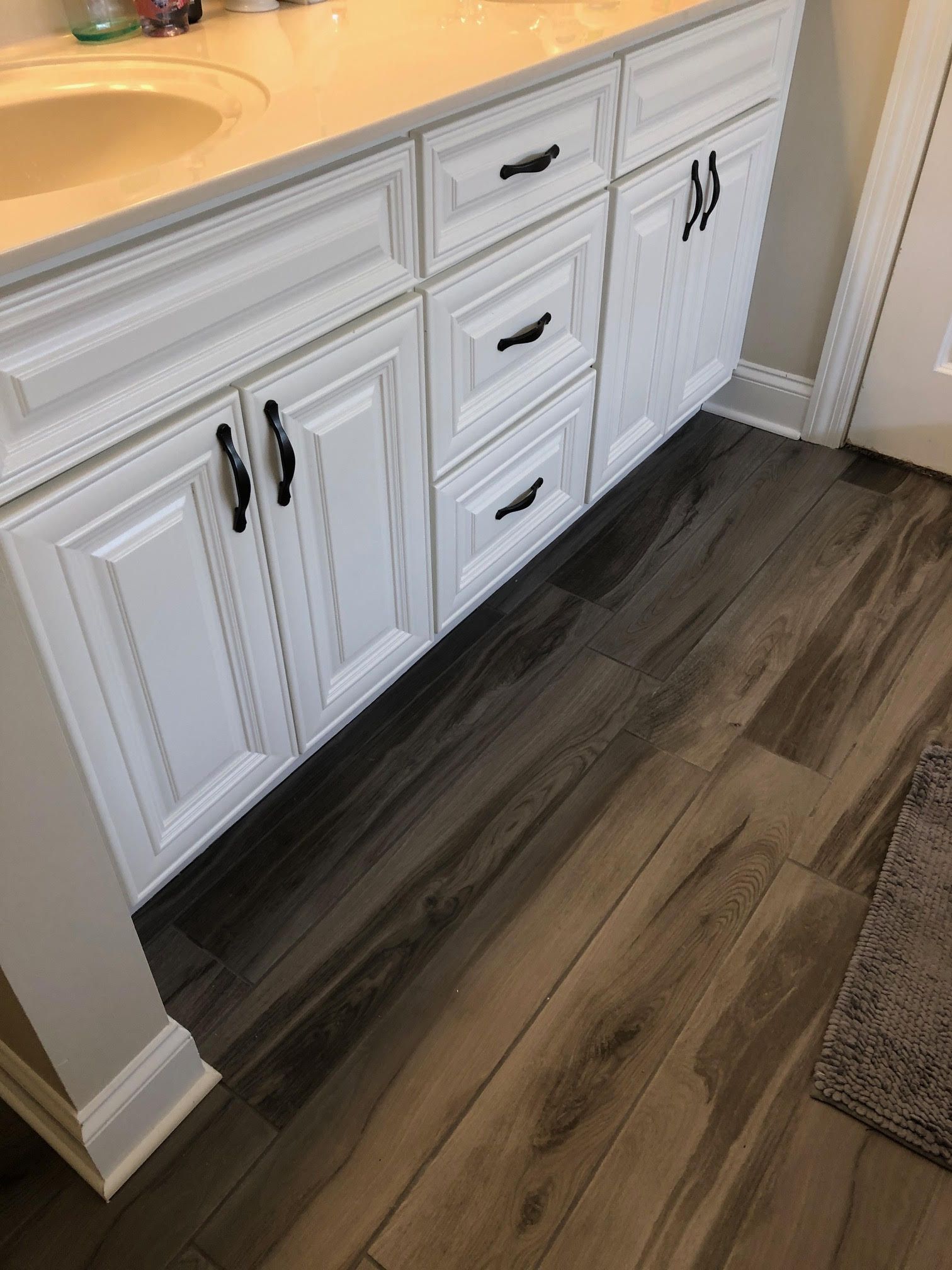 White bathroom vanity with black hardware, on wood-look tile floor.
