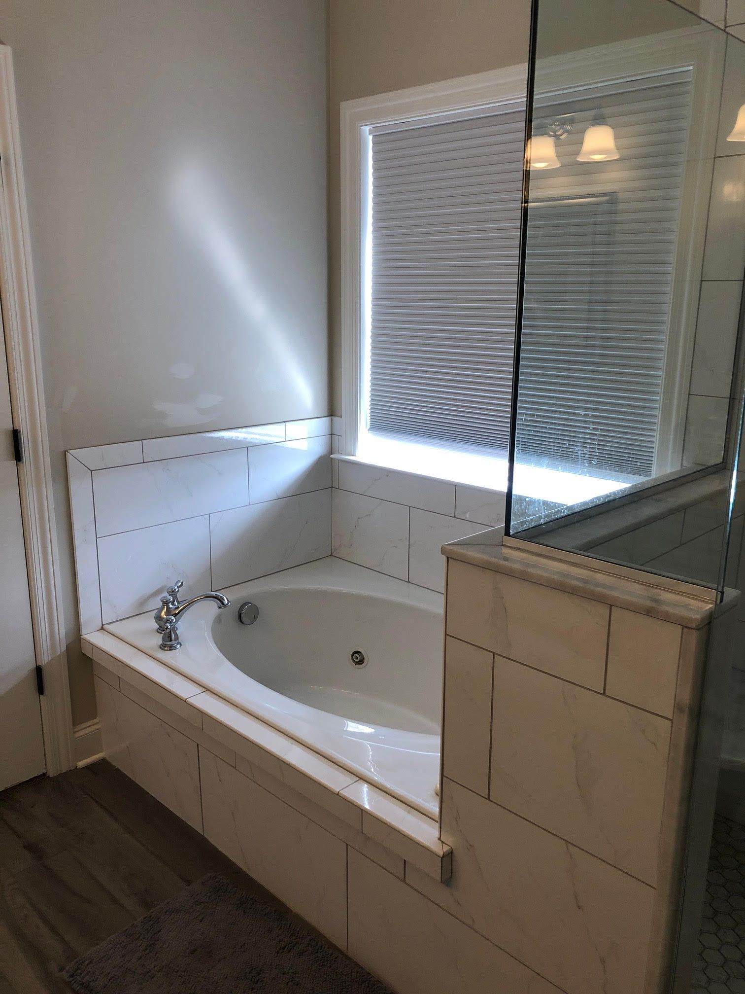 Bathroom with a white tiled jacuzzi tub under a window with blinds. A glass shower is visible.
