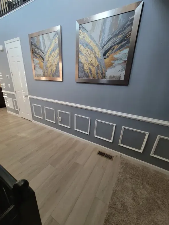 A blue wall with two framed artworks above a white molding, next to a door and light wood floor.