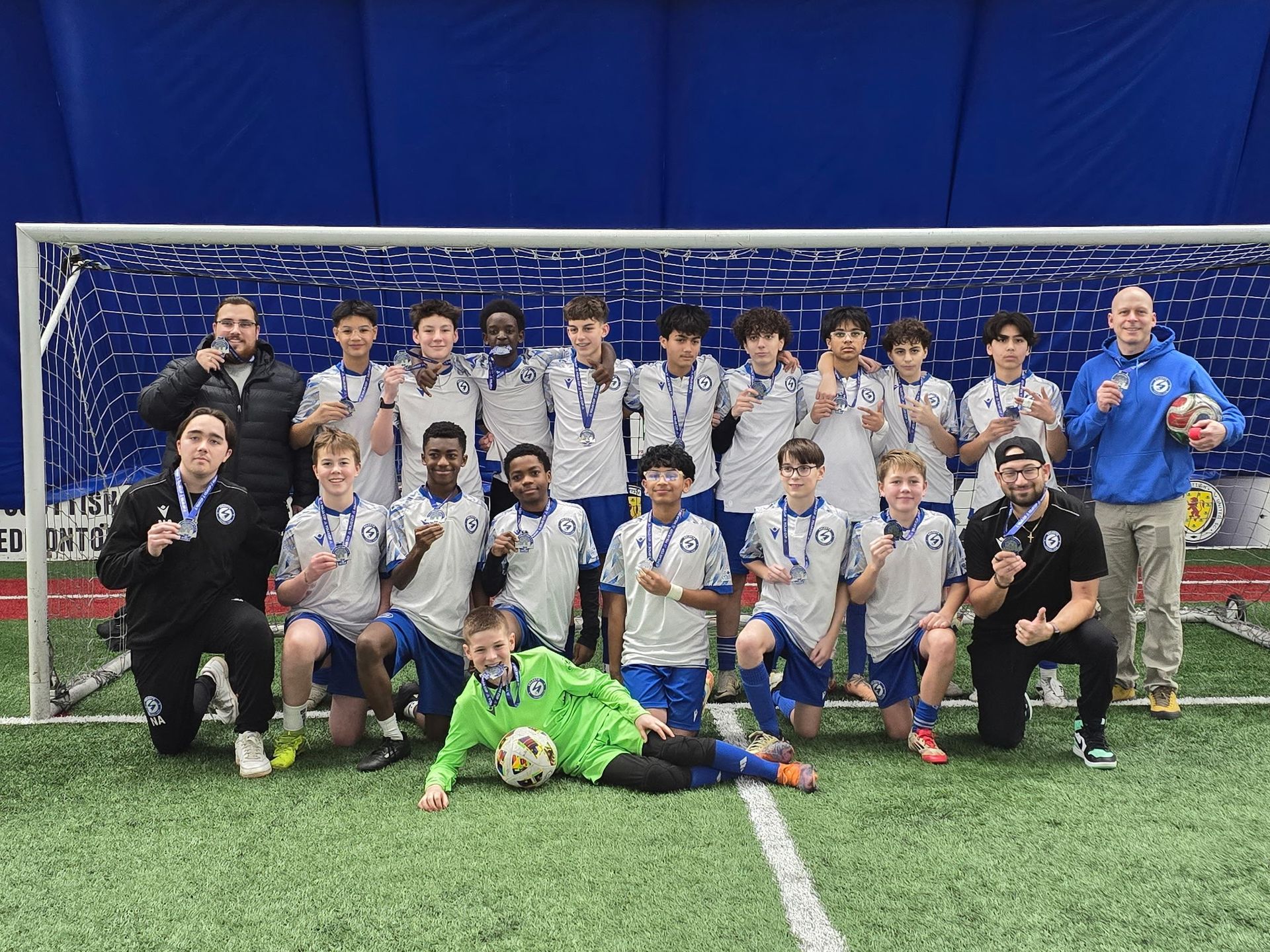 A soccer team and coaches pose in front of a blue indoor goal, wearing medals and team uniforms on an artificial field.