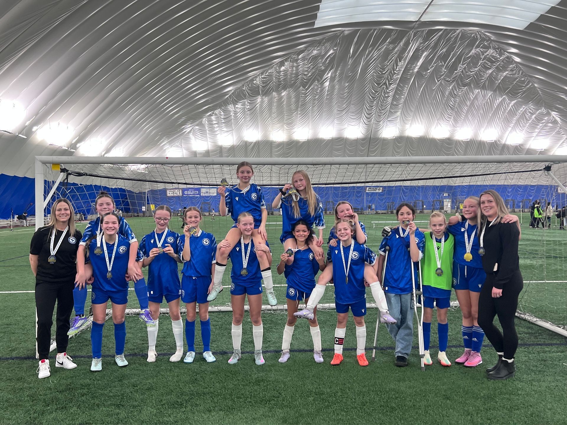 A youth soccer team in blue uniforms and medals stands on an indoor turf field in front of a goal net.