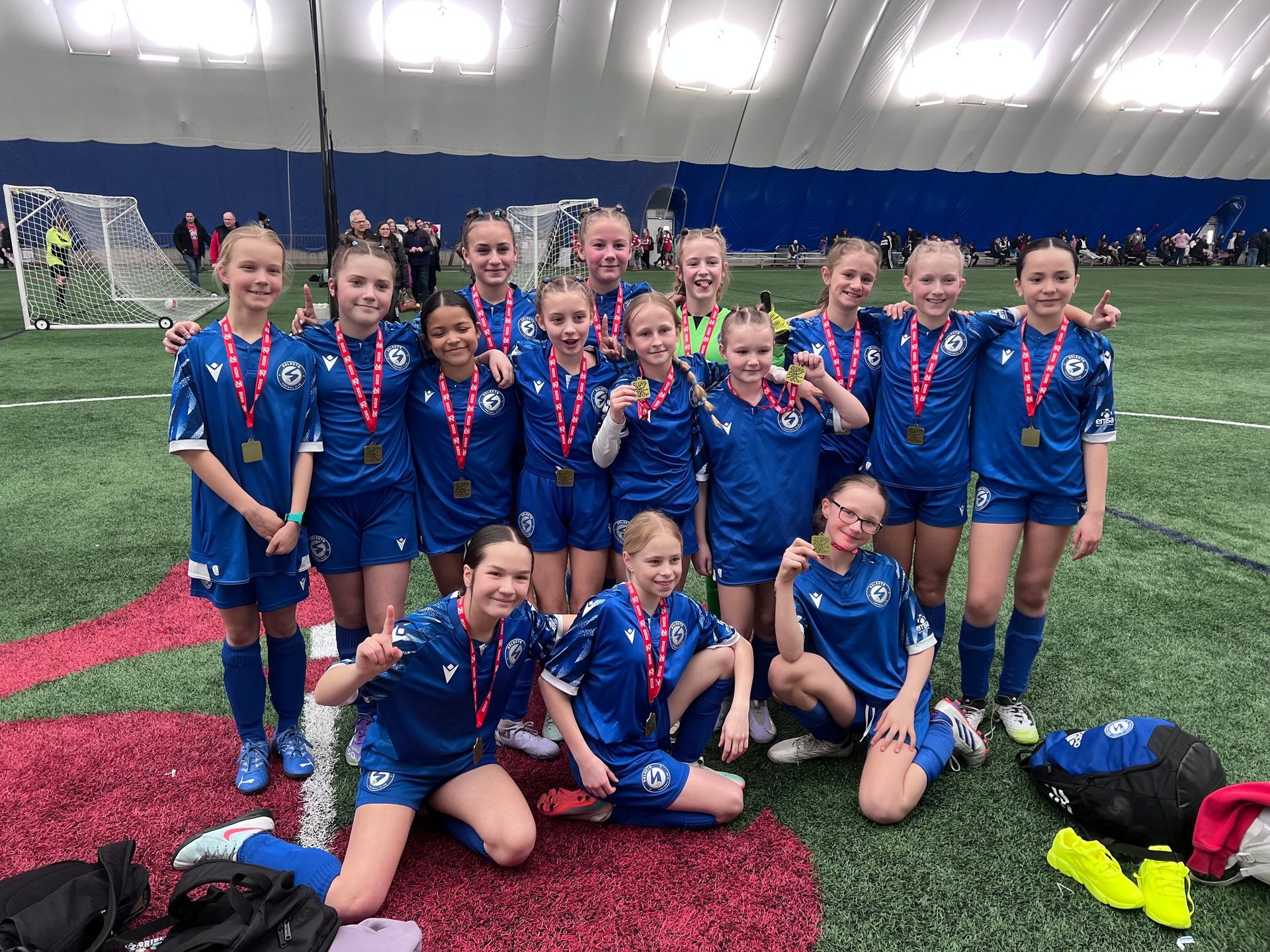A youth soccer team in blue uniforms and medals poses for a photo on an indoor turf field.