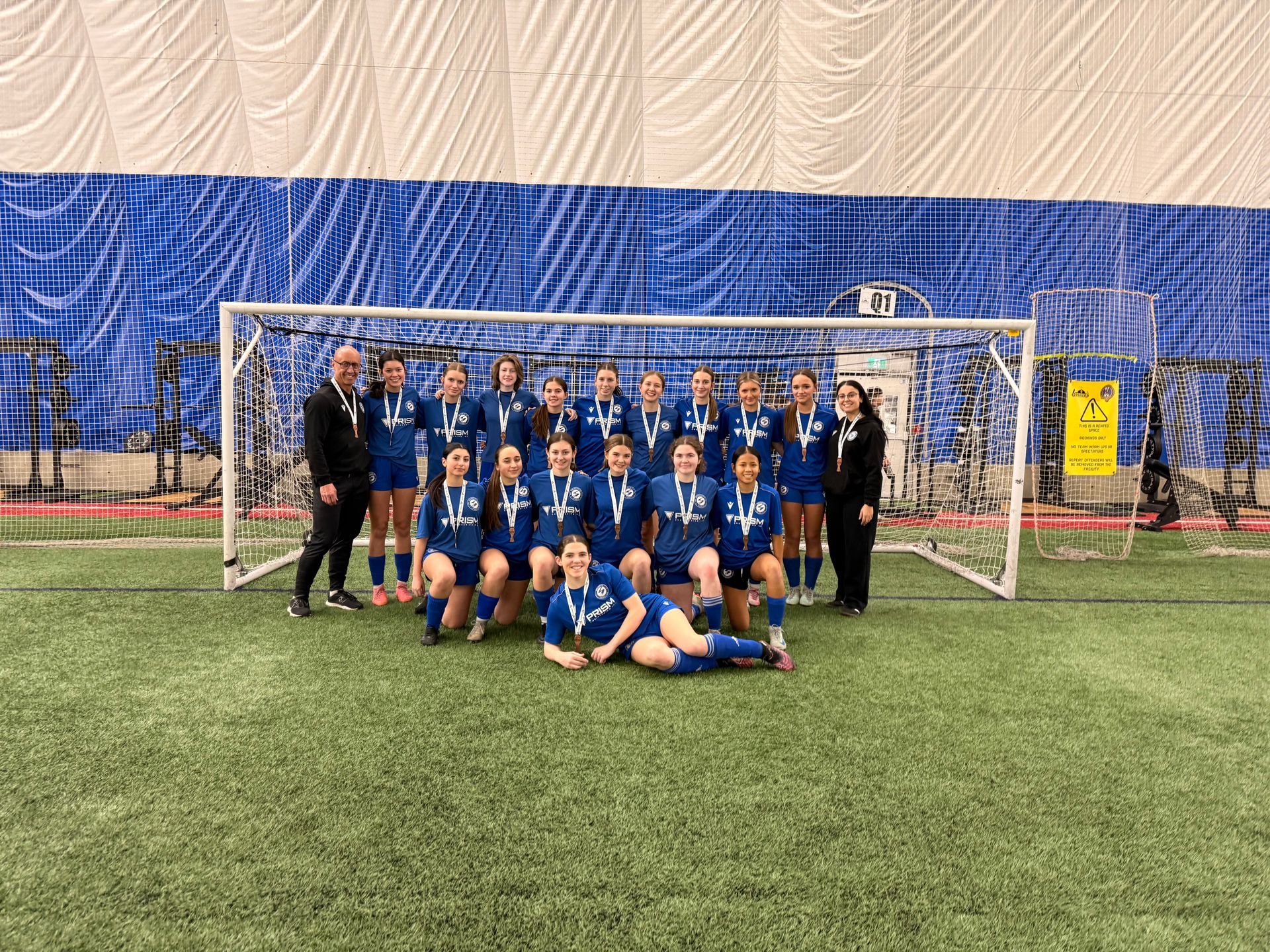 A youth soccer team in blue uniforms posing for a photo with two coaches in front of a goal on an indoor turf field.
