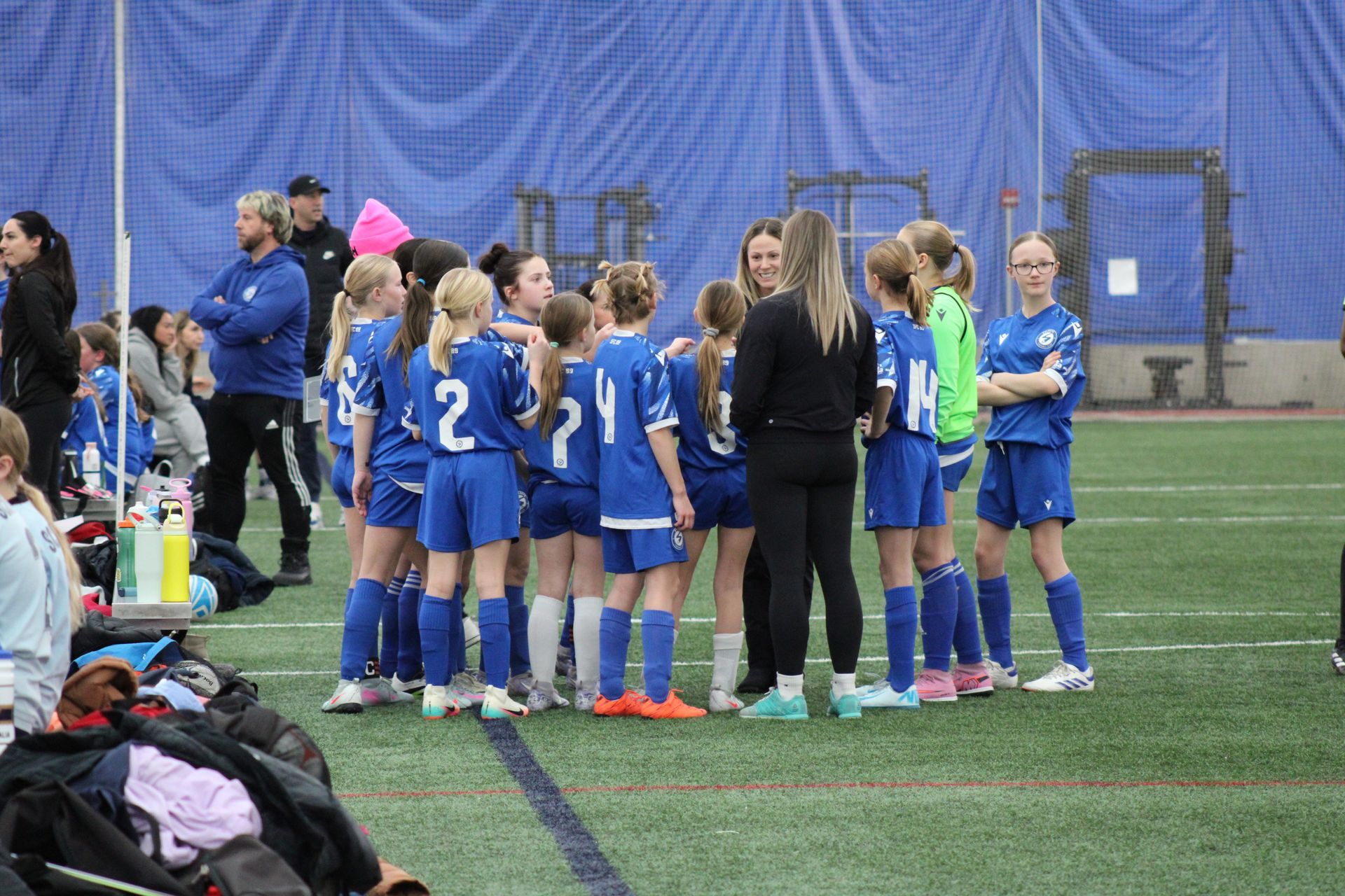 A youth soccer team in blue uniforms huddles with coaches on an indoor turf field.