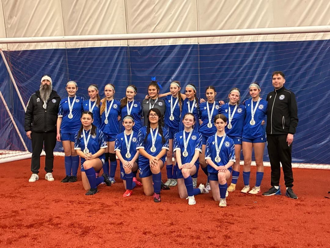 A youth soccer team in blue uniforms and medals stands on an indoor turf field in front of a blue and white net.