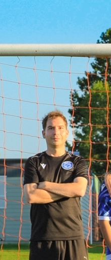 A coach with arms crossed, wearing a black shirt with a blue emblem, stands in front of a soccer goal net.
