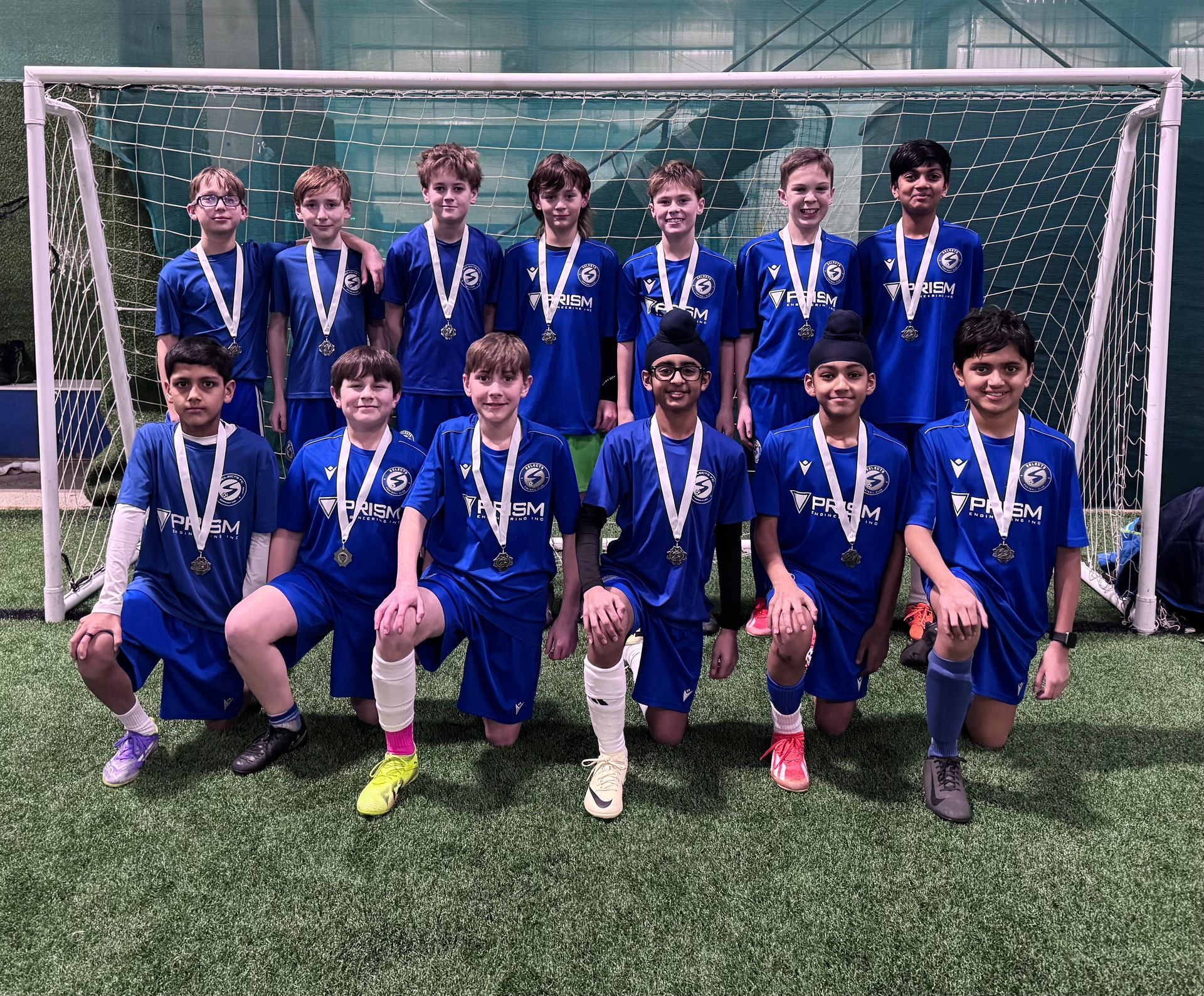A soccer team in blue uniforms stands in front of a goal net, wearing medals after a tournament.