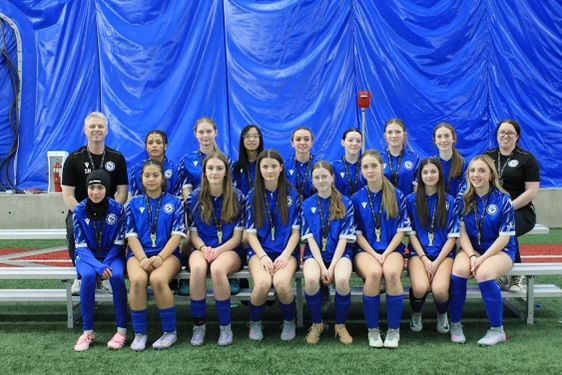 A team of soccer players in blue jerseys and two coaches posing for a group photo in an indoor sports facility.